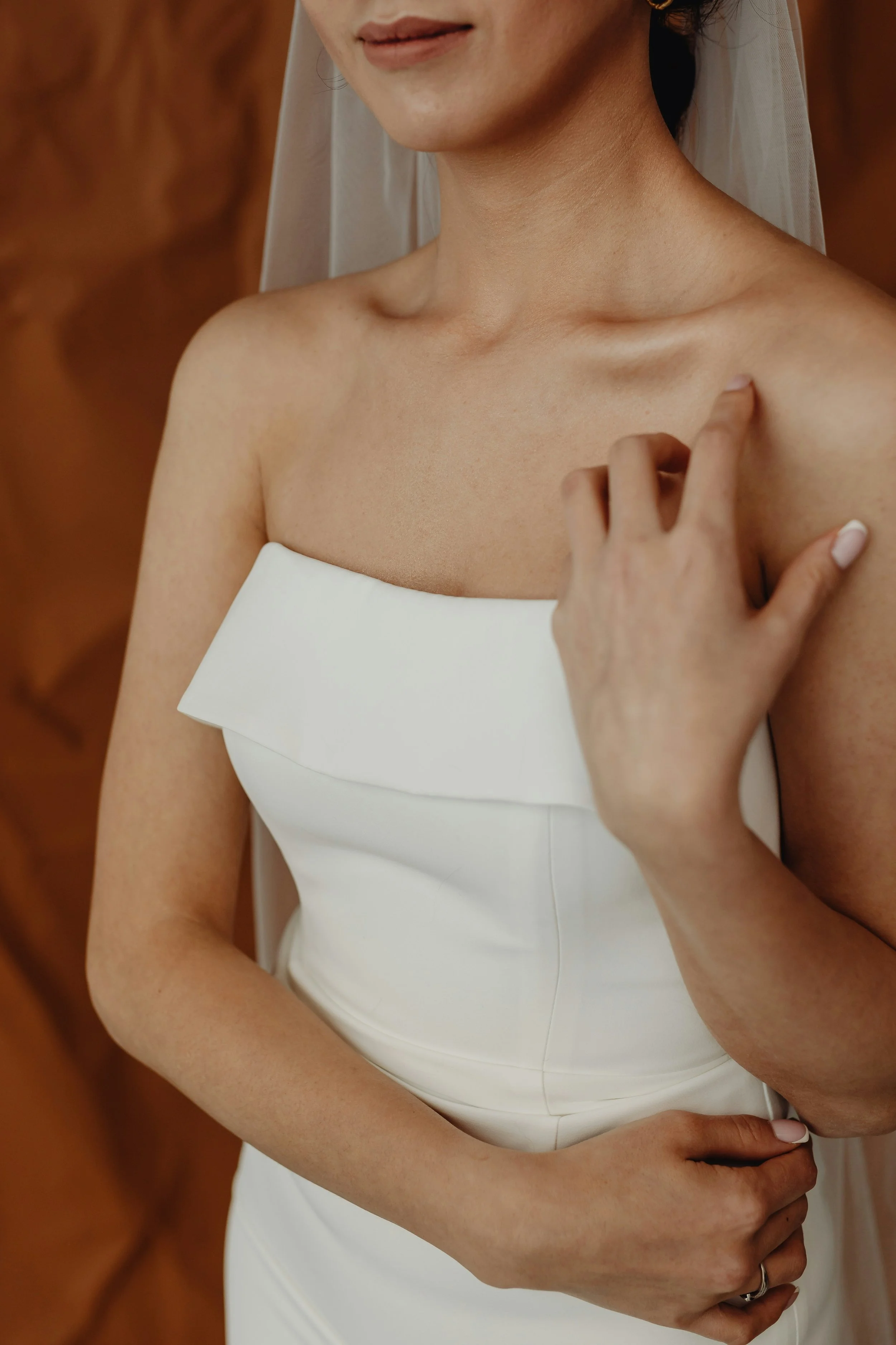 Close-up of a woman in a white wedding dress, touching her shoulder with one hand and resting her other hand on her waist, with a blurred background.