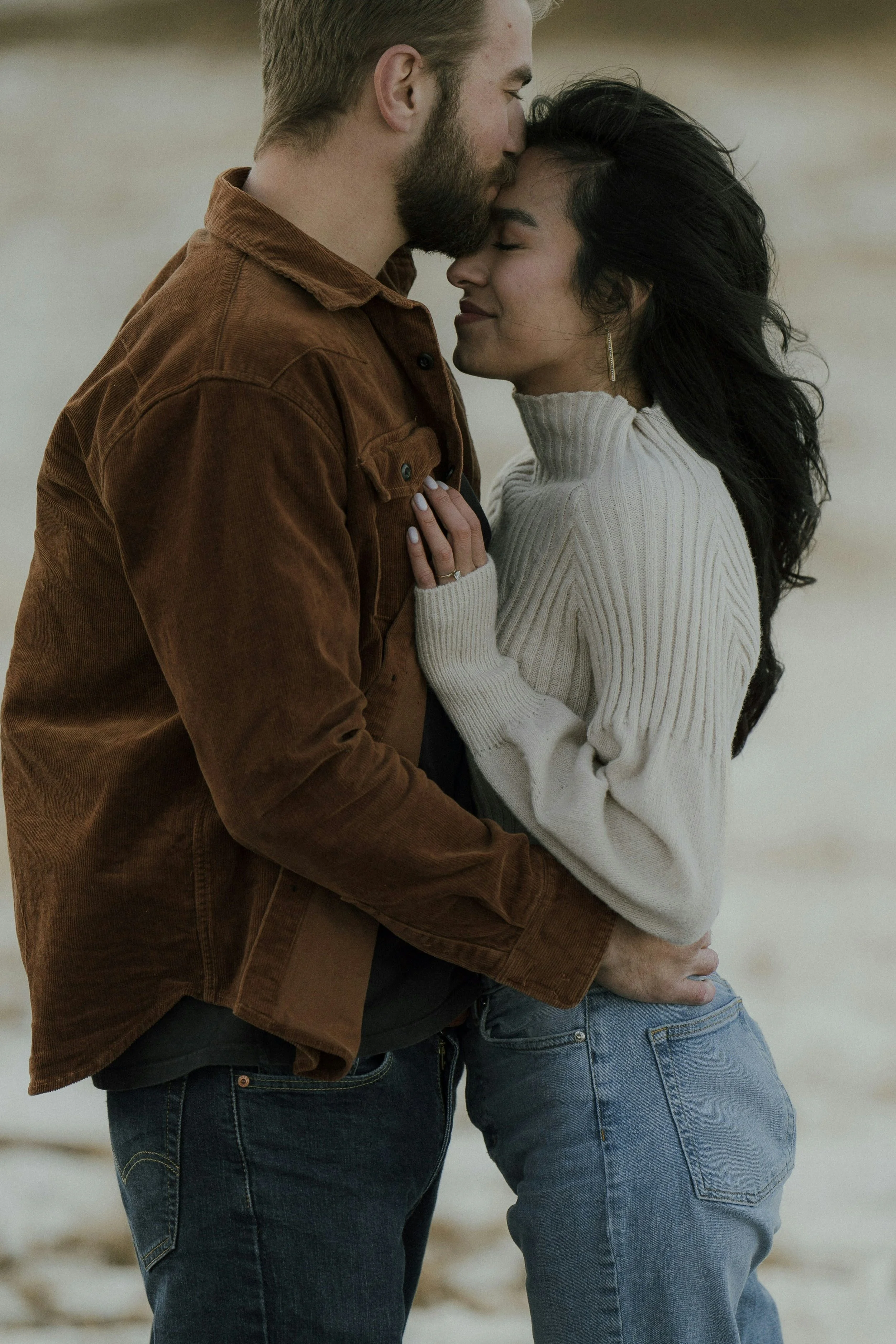 A man and woman embrace each other closely, with the man kissing the woman's forehead on a beach.