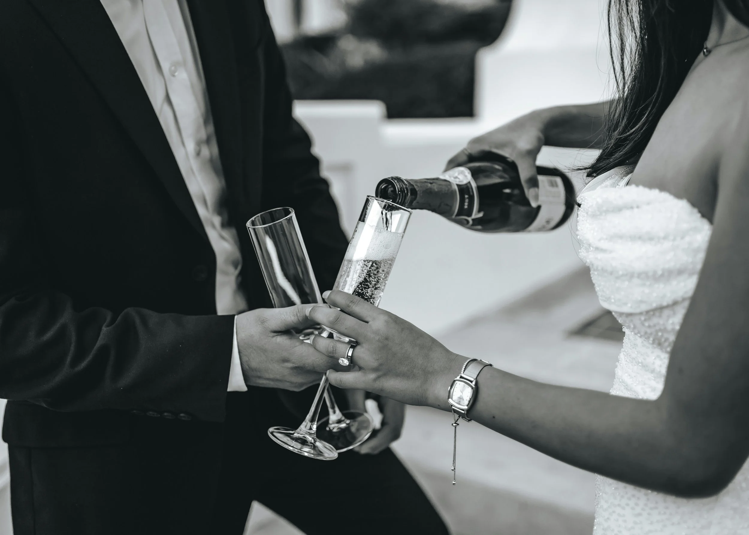A woman in a wedding dress being handed a bottle of champagne by a person in a suit, while she pours champagne into her glass.