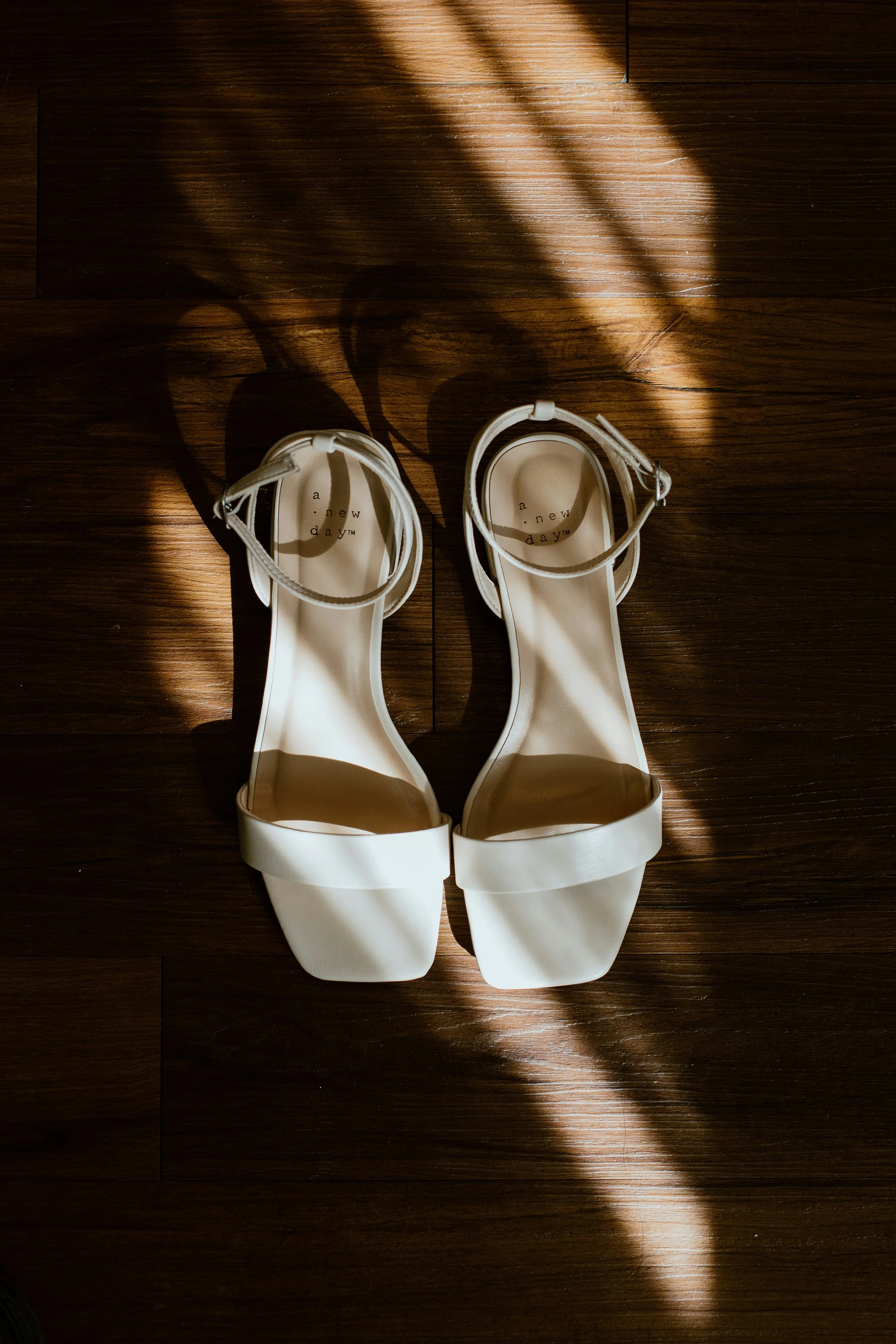 White sandals with ankle straps and open toes on a wooden floor, illuminated by sunlight creating a shadow pattern.