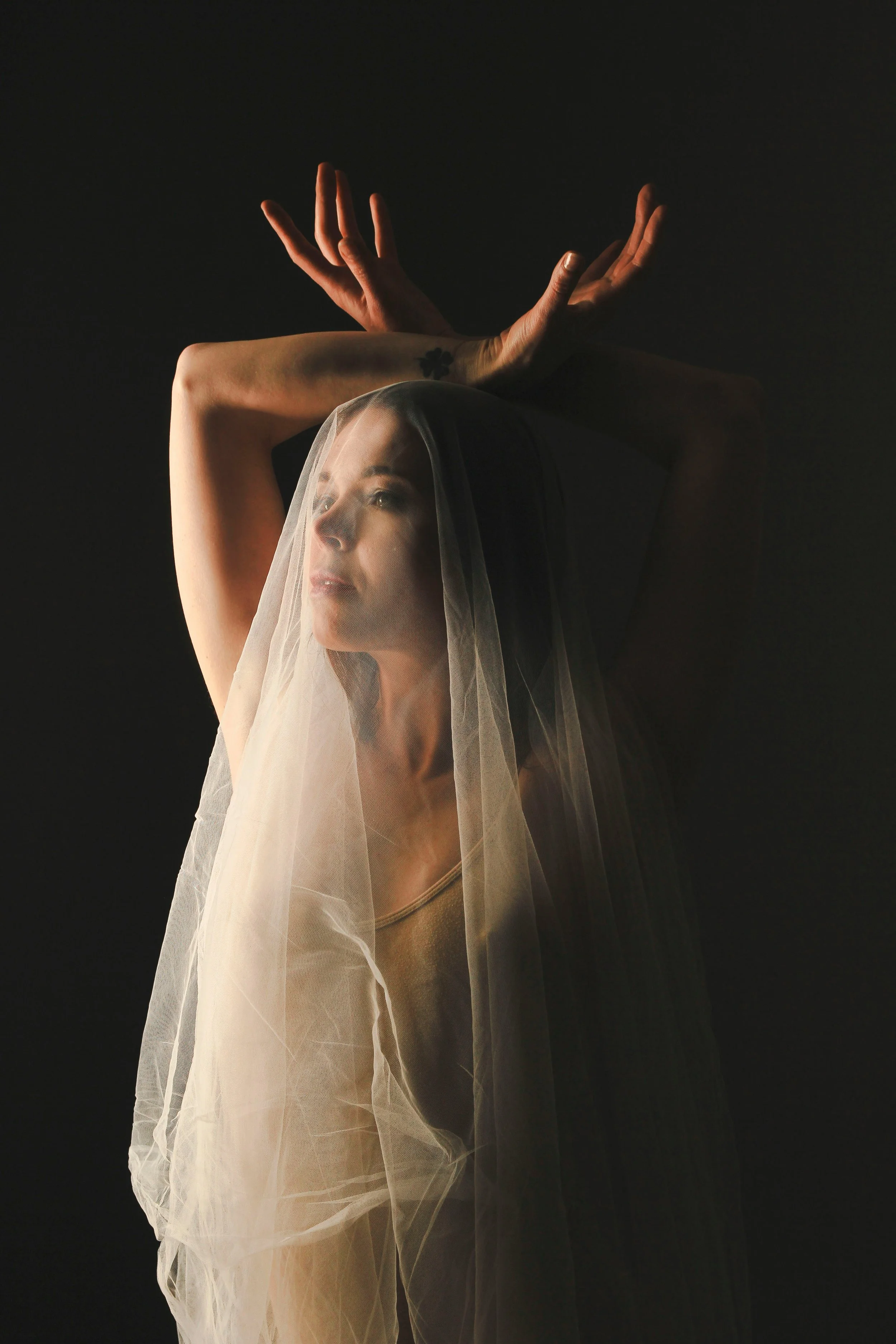A woman with a veil over her face, looking contemplative, with her arms raised above her head against a dark background.