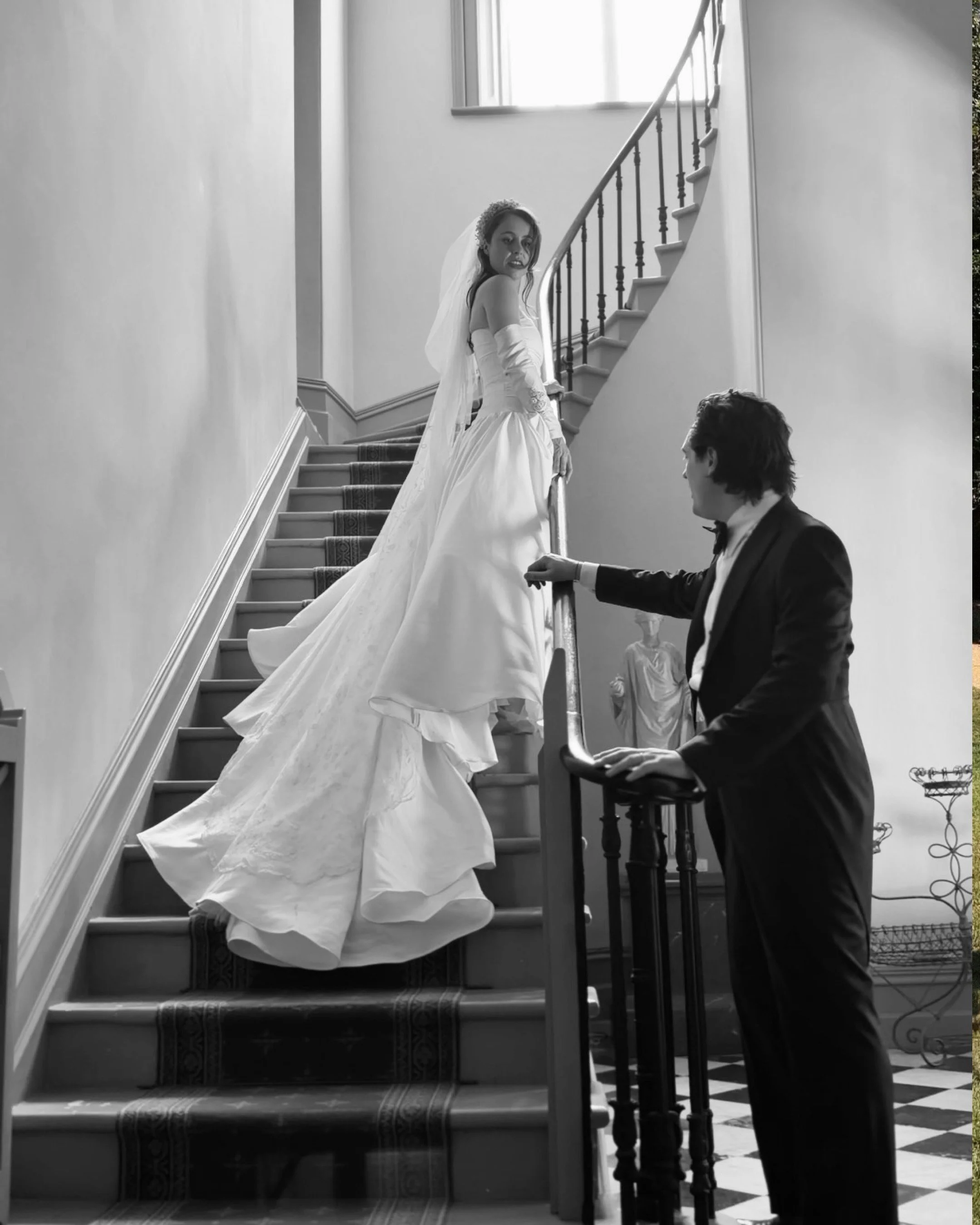 A woman in a wedding dress and veil standing on a staircase, looking back at a man kneeling in front of her, holding her hand, in a black and white photograph.