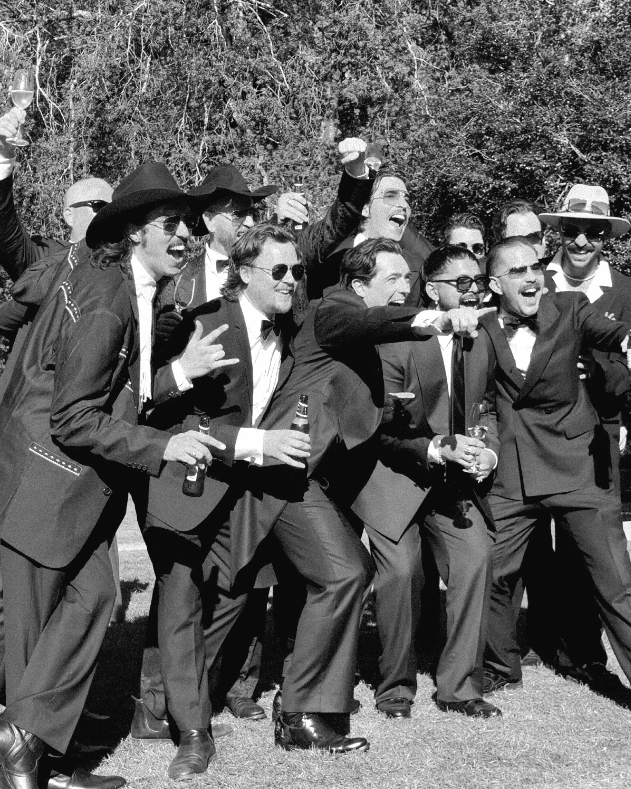 A group of men dressed in tuxedos, some wearing sunglasses and bow ties, running outdoors in a joyful manner, holding drinks, with trees in the background.