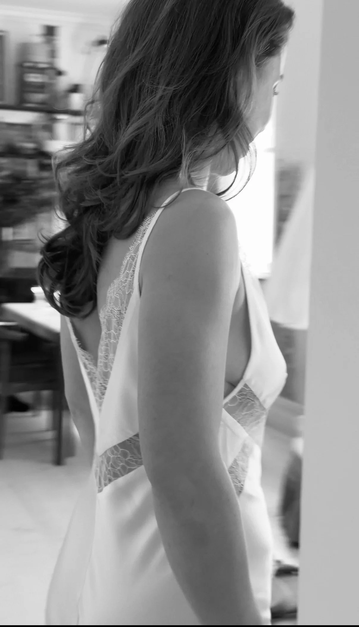 A woman with curly shoulder-length hair wearing a satin and lace nightgown, standing indoors, seen in profile.