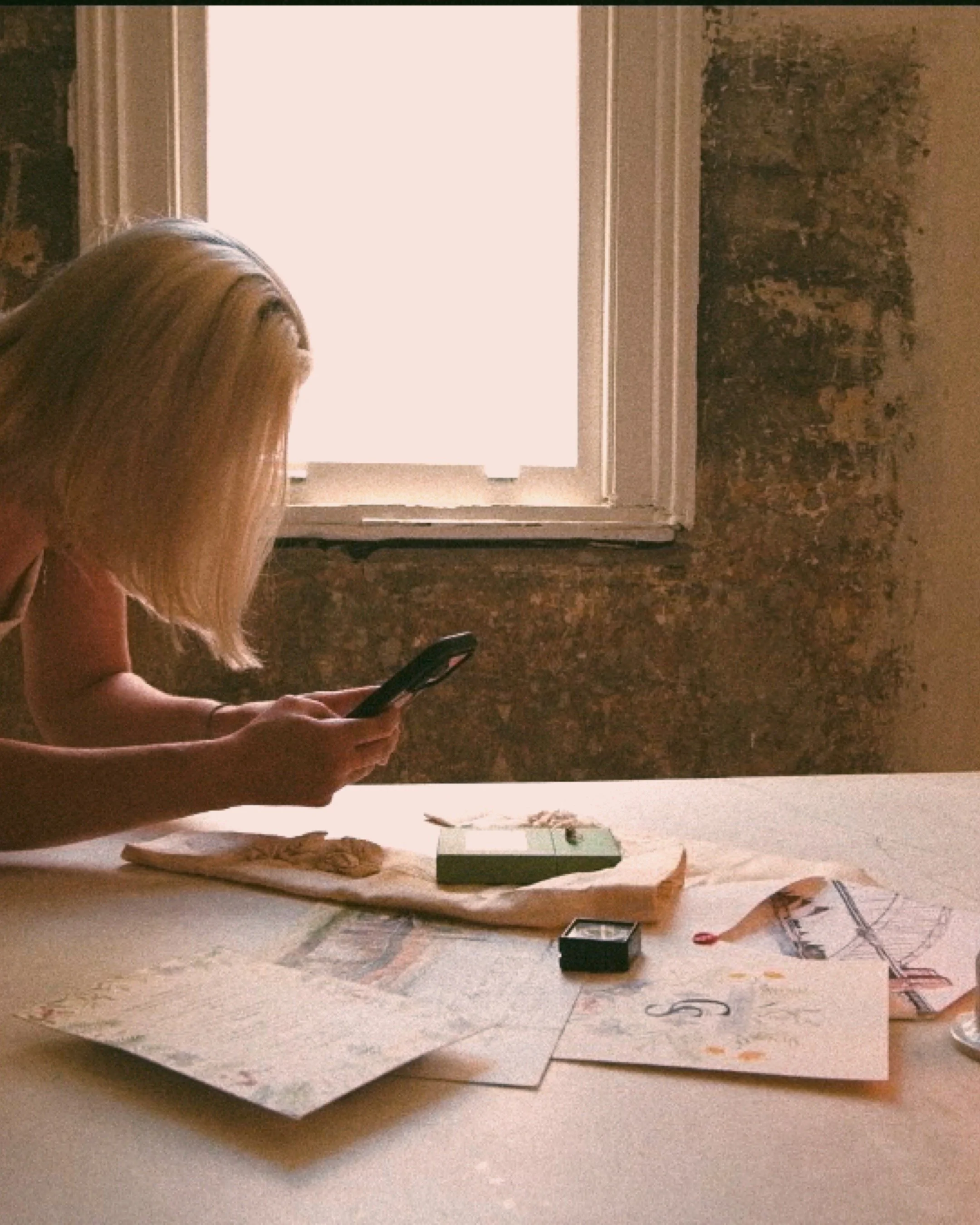 A person with long blonde hair is sitting on the floor, using a smartphone, in front of a window with light coming through. The floor is covered with papers, a small green and black box, and various art supplies like scissors and markers.