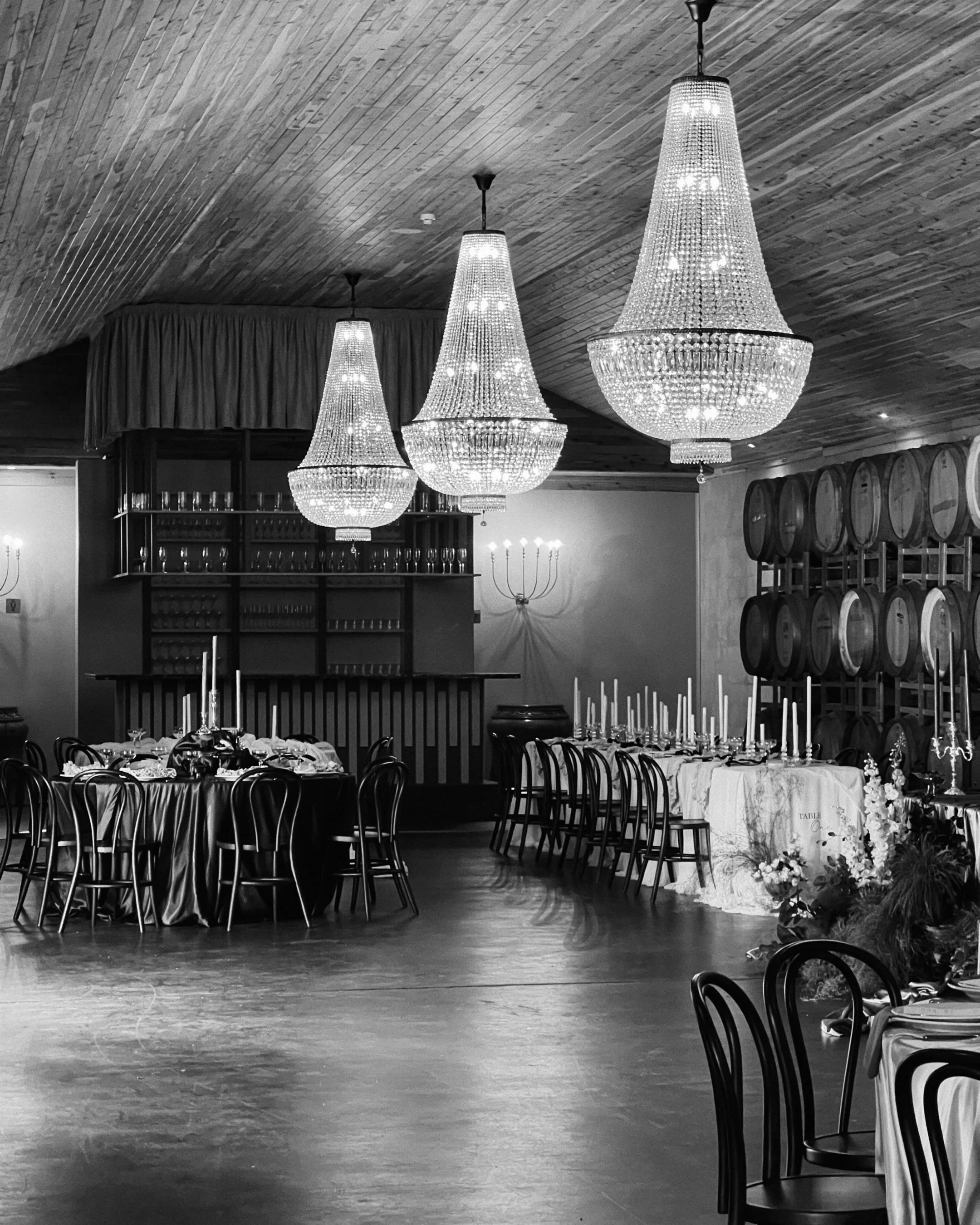 Elegant dining area with multiple chandeliers hanging from a wooden ceiling, tables set with white tablecloths, candles, and floral arrangements, and wine barrels lining the wall.
