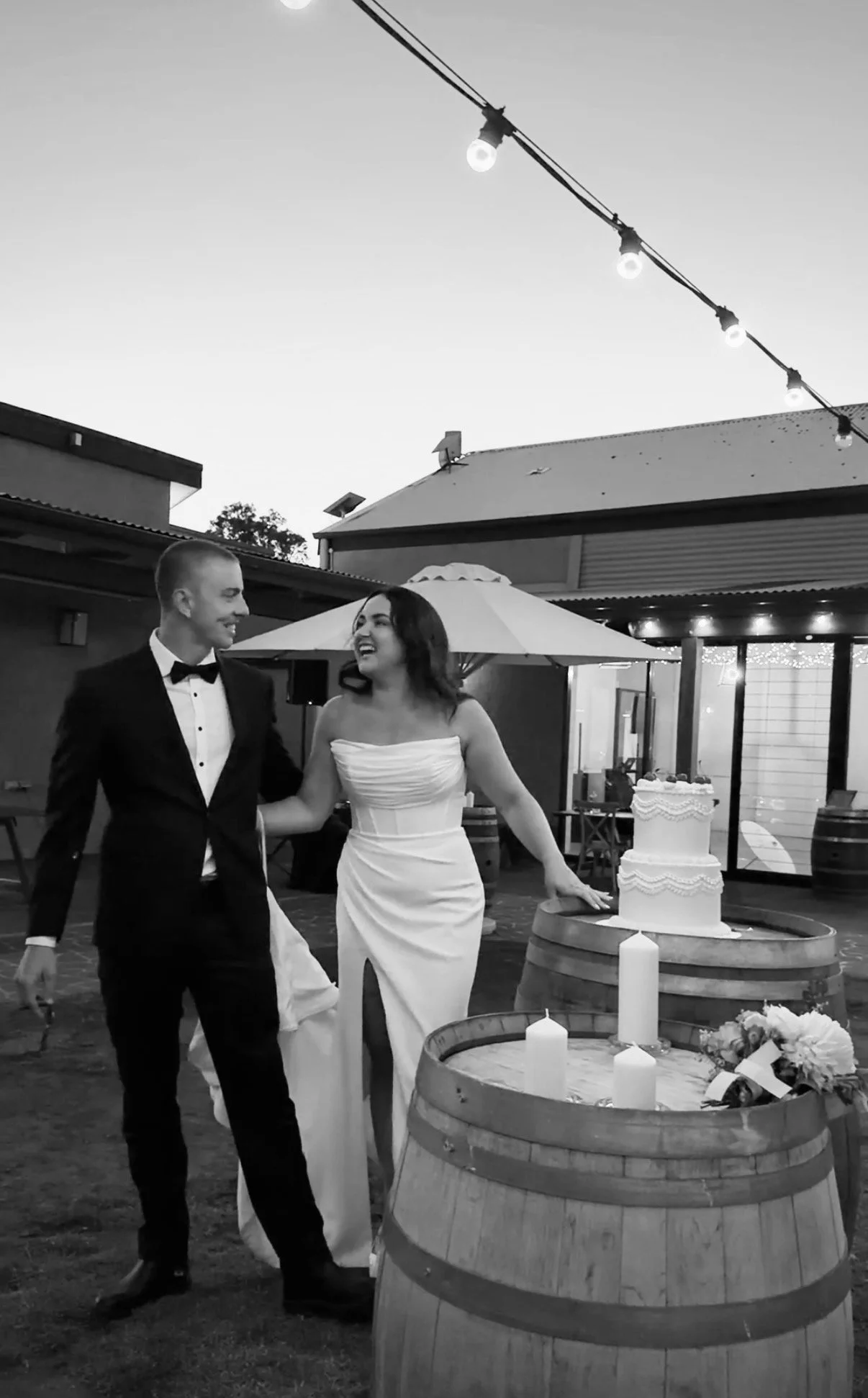 A couple in wedding attire holding hands and smiling at each other outdoors in the evening next to a wedding cake on a table decorated with candles.