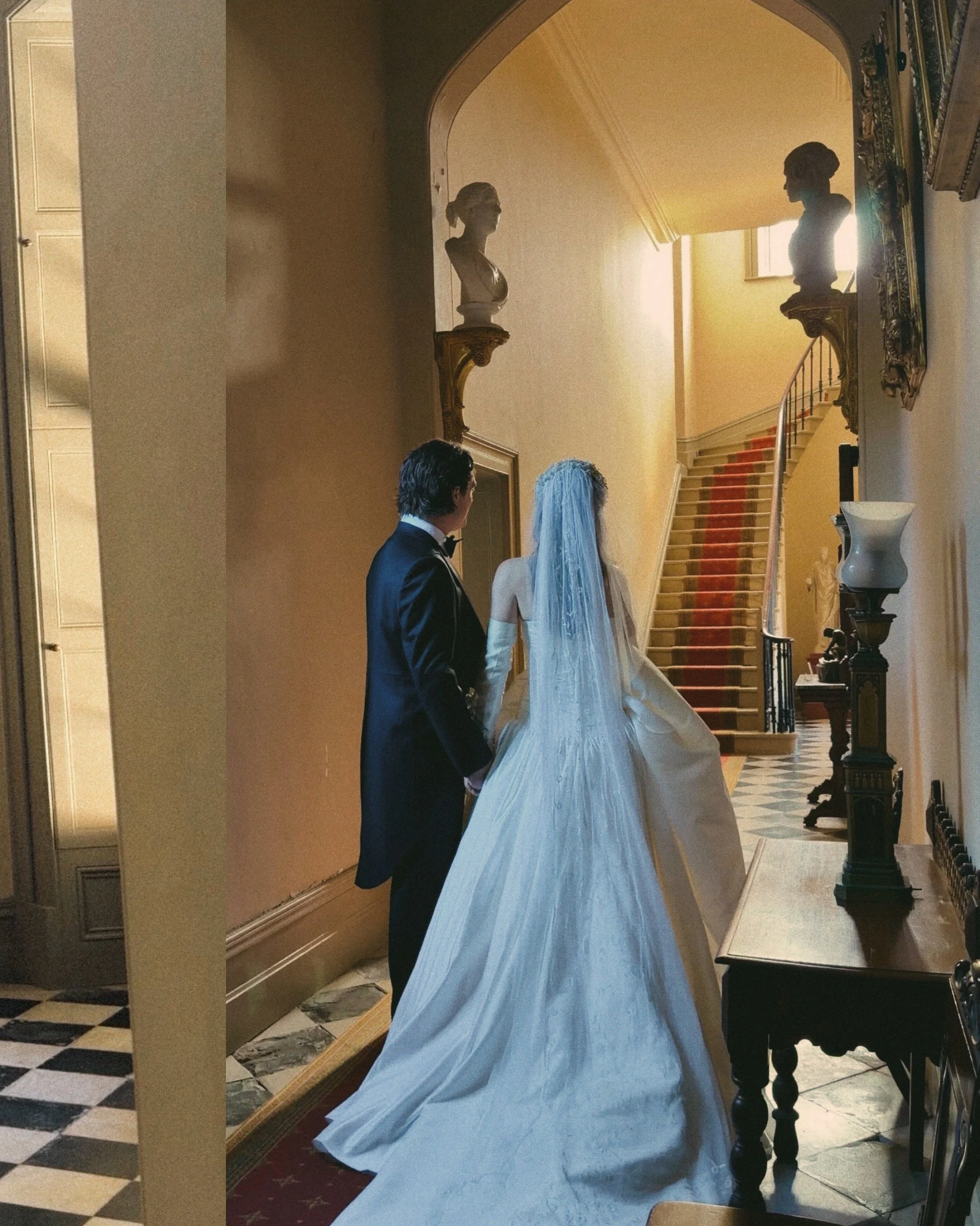 Inside a grand house with a staircase, two classical bust sculptures displayed on ornate wall shelves, and a bride with white dress and veil standing next to a man in a tuxedo, possibly during a wedding.