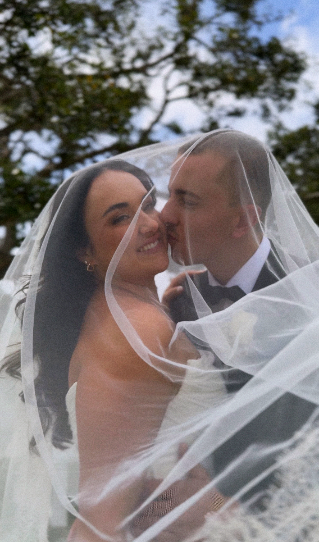 A newlywed couple, the bride with dark hair and the groom with short hair, smiles and kisses under a wedding veil outdoors with trees and sky in the background.