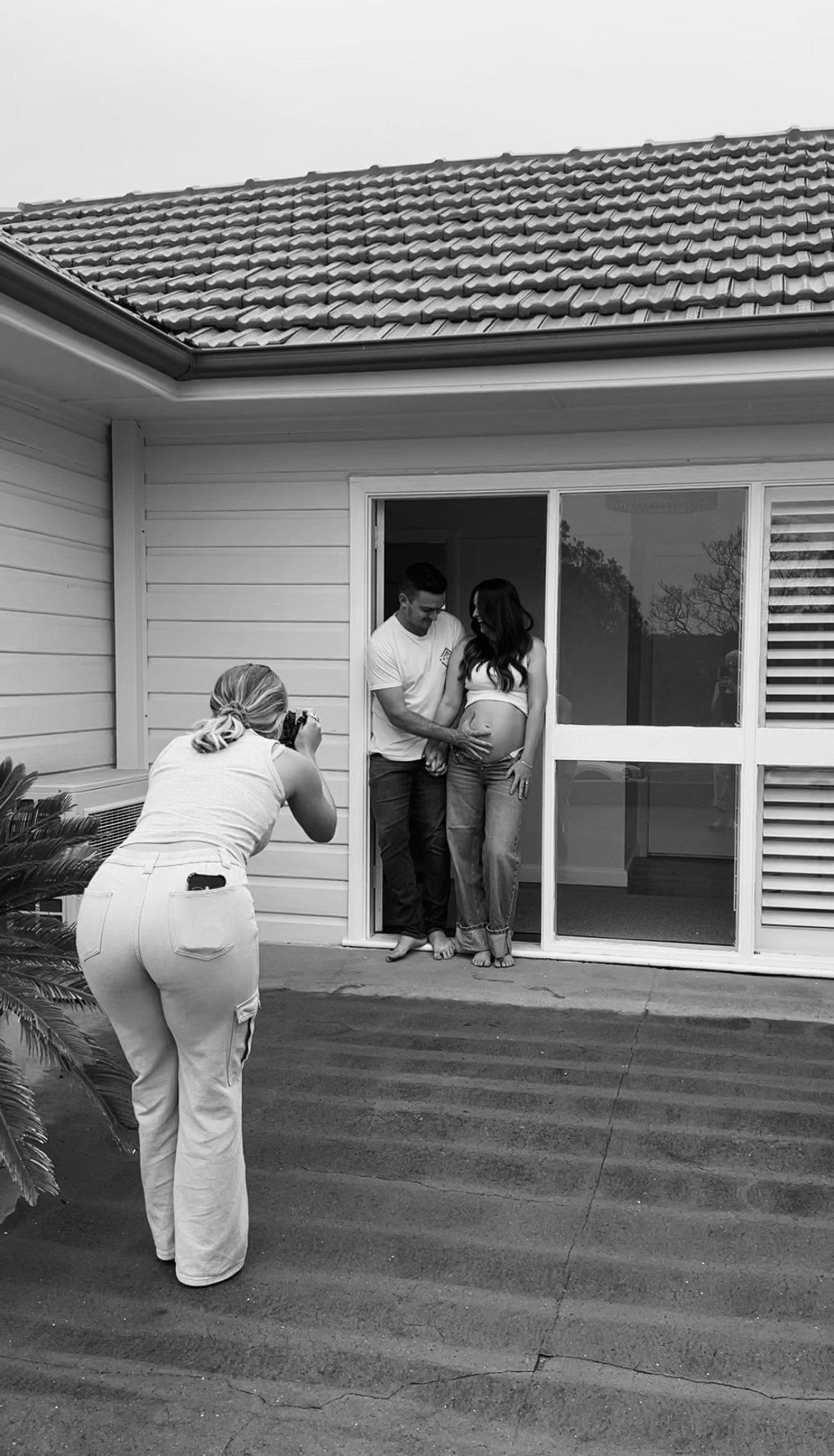 A pregnant woman with a man in front of a house, while a woman takes a photo outside the house.