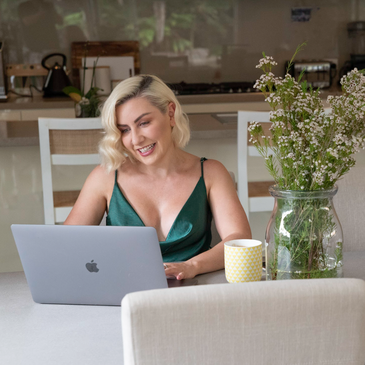 A woman with blonde hair sitting at a table using a silver MacBook laptop, smiling, with a vase of white flowers and a yellow mug nearby, in a cozy kitchen setting.