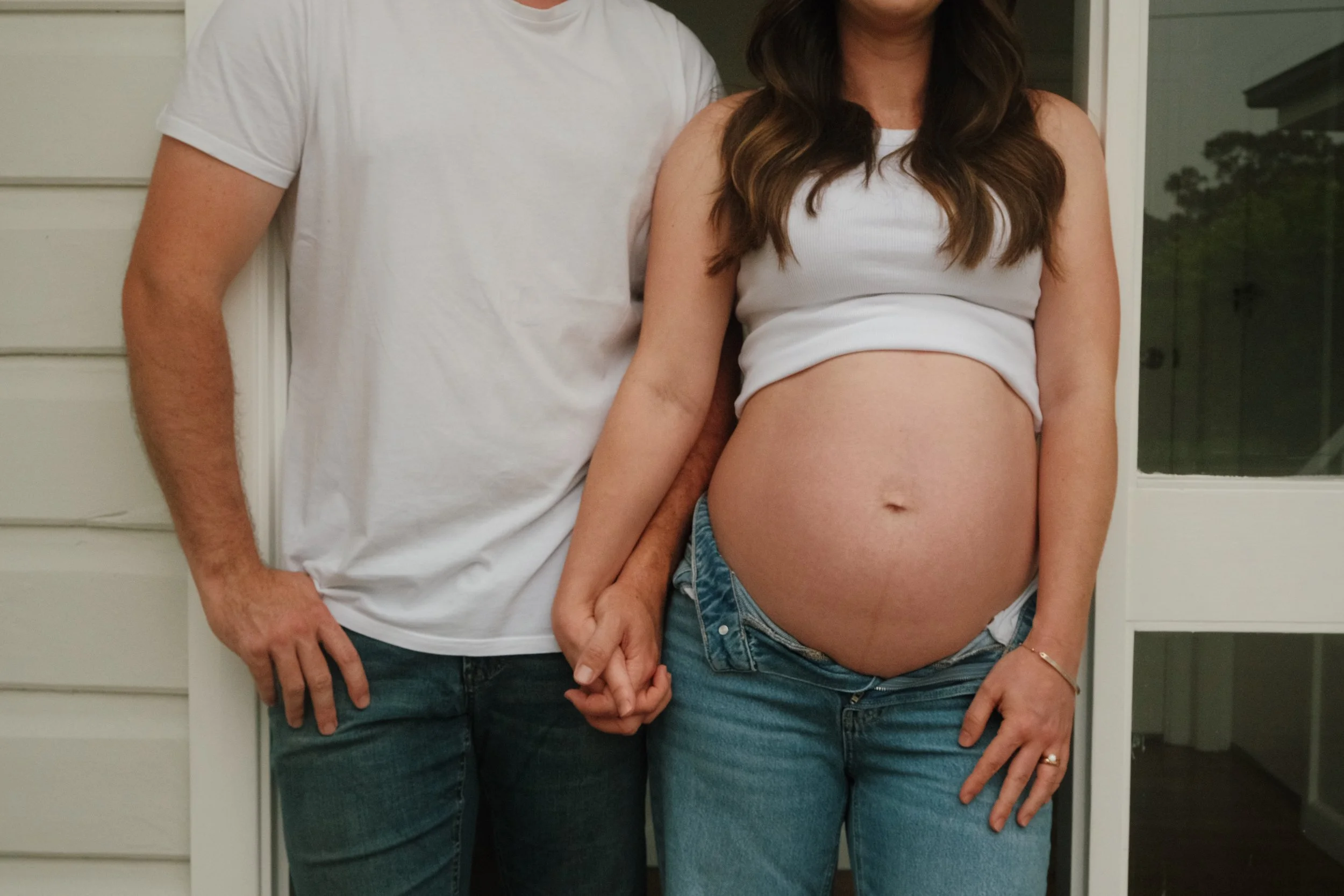 A pregnant woman holding hands with a man, both standing outside near a door, with the woman showing her exposed belly.