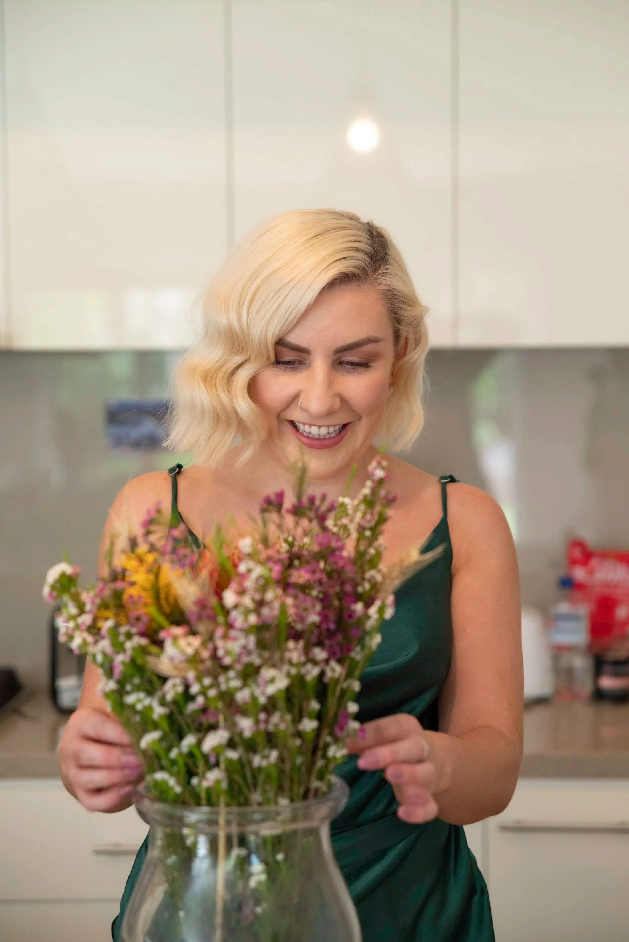 A woman with platinum blonde hair styled in loose waves, smiling as she looks at a bouquet of colorful flowers in a glass vase.