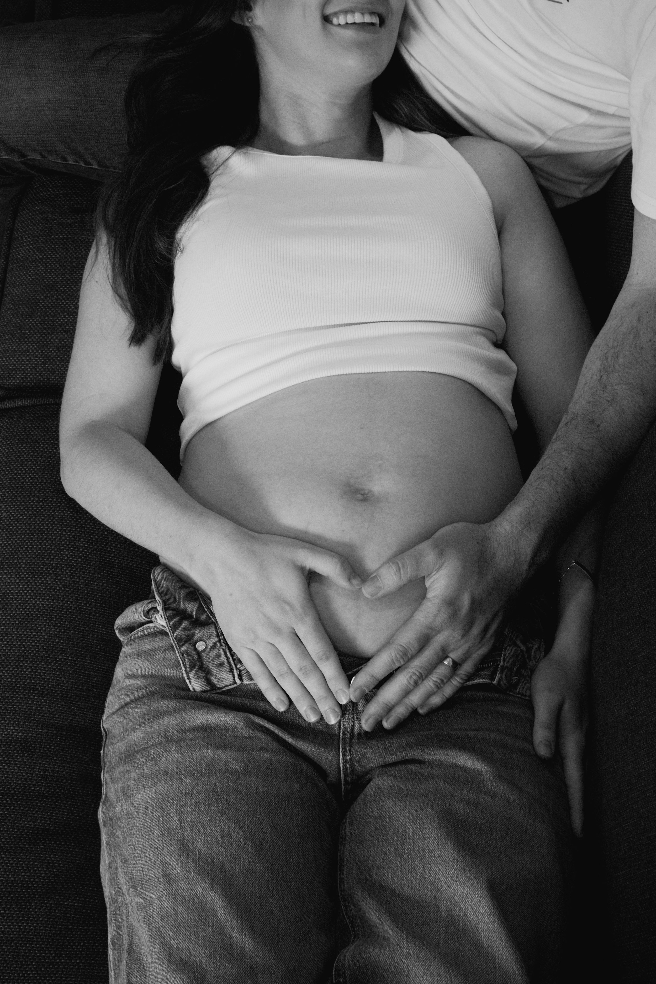 A pregnant woman lying on a couch, smiling, with a man's hands gently resting on her belly.