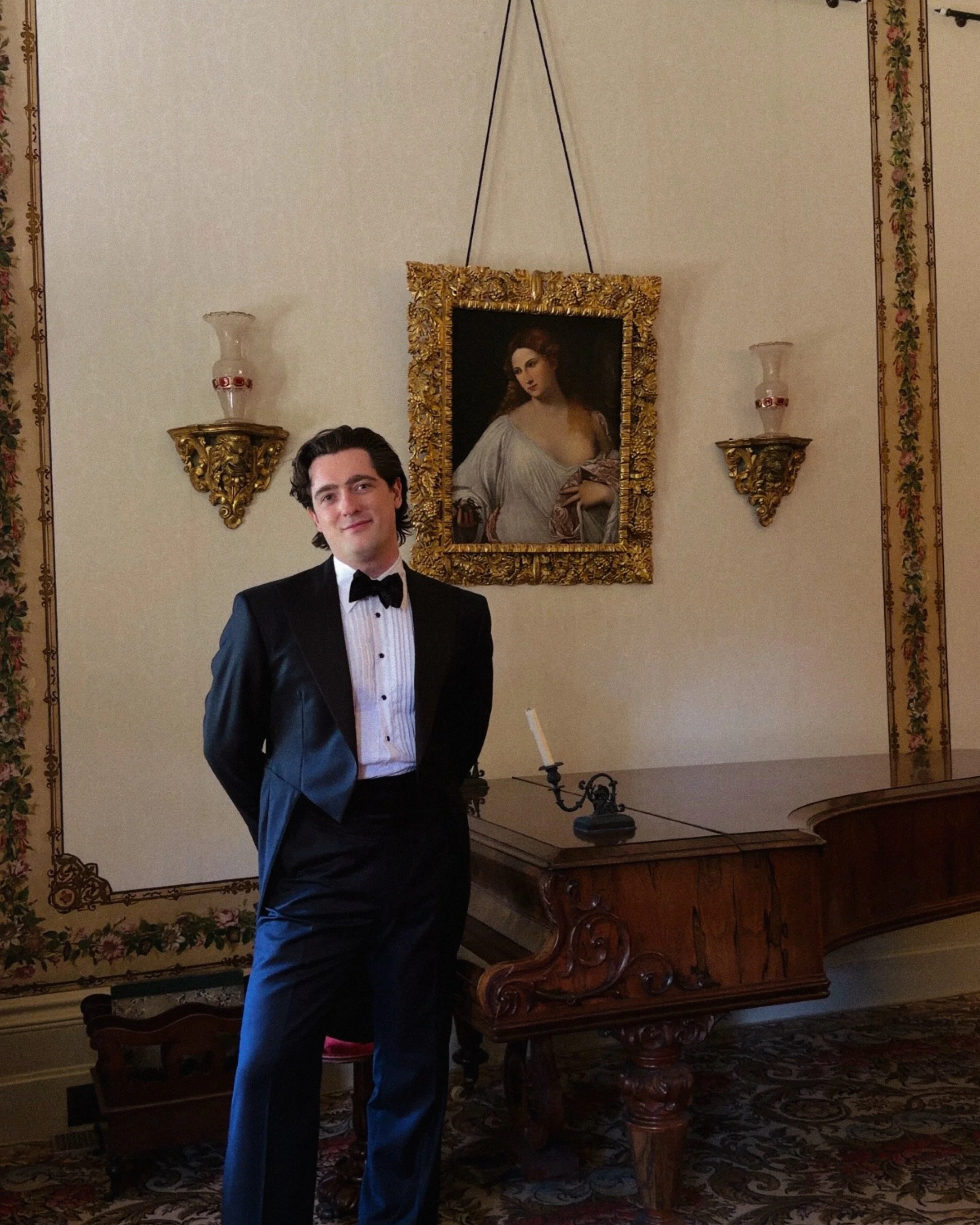 A man in a tuxedo standing indoors with elegant decor, including a gilt-framed portrait of a woman on the wall and ornate wall sconces.
