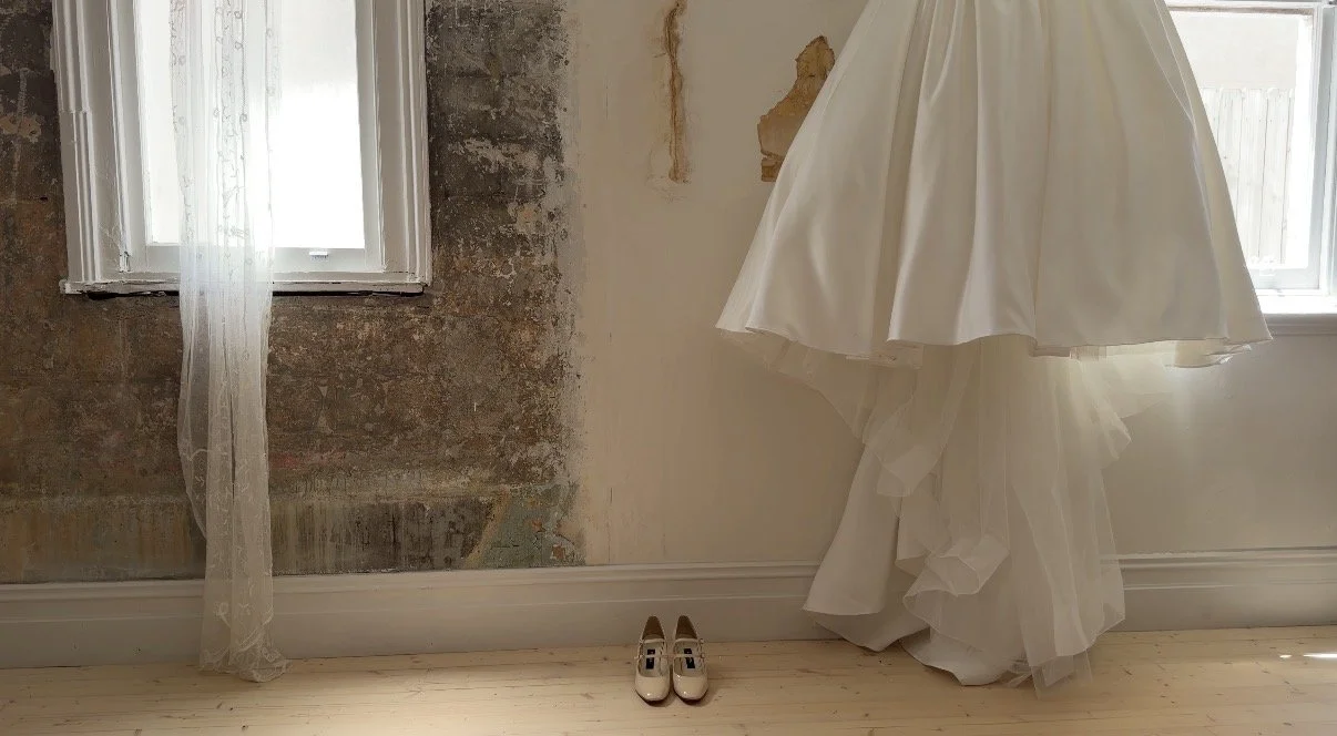 A white wedding dress hanging in a room with two windows, one of which has sheer curtains. A pair of white shoes are placed on the floor beneath the dress, and the room has a partially exposed wall with peeling paint.