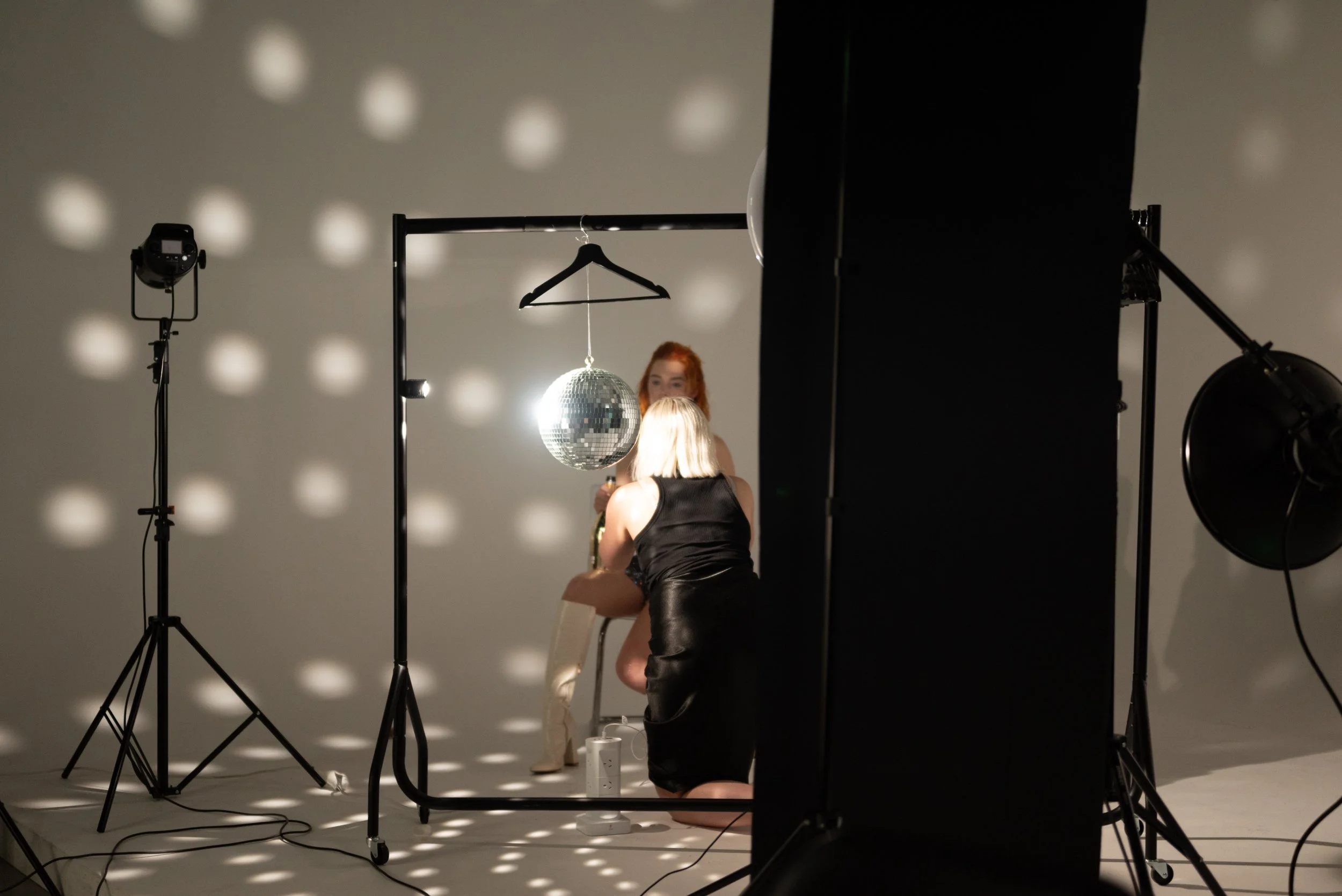 Photo of a photoshoot setup with studio lighting, a disco ball, and two women, one with blonde hair and the other with red hair, in front of a backdrop with light spots.