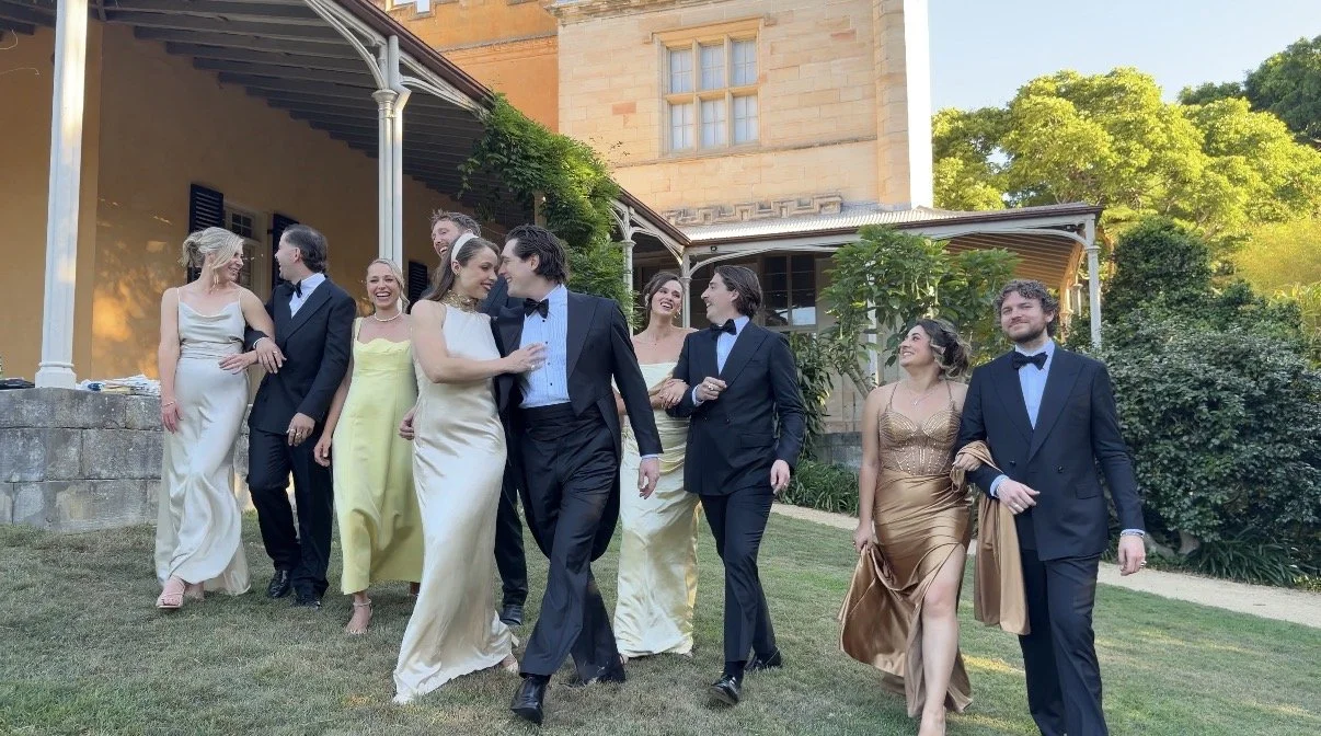 Group of people in formal attire walking on grass outside a historic building, smiling and enjoying each other's company.