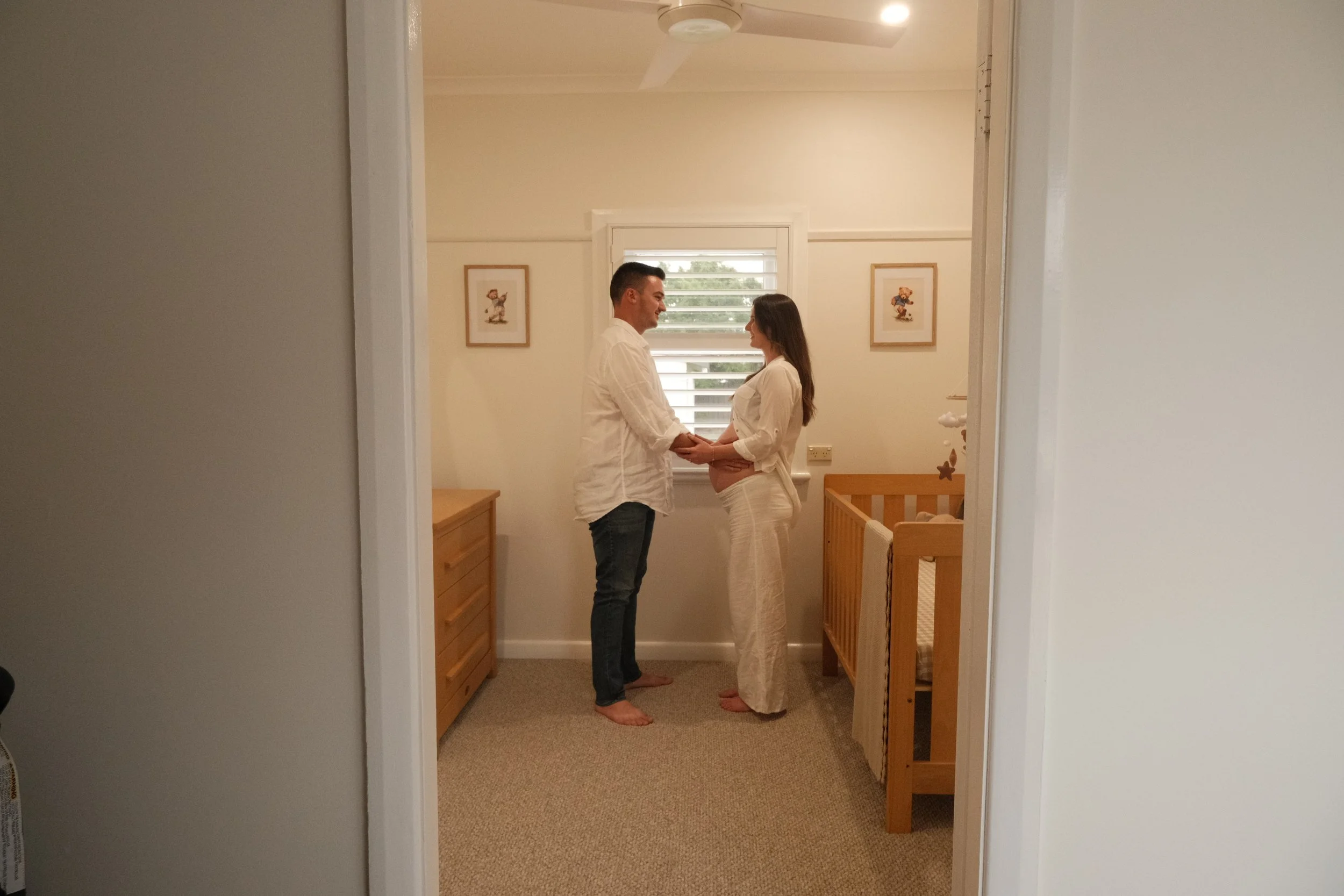 A couple standing in a baby's nursery, facing each other and holding hands. The woman is pregnant and wearing white pants and a white blouse, while the man is dressed in a white shirt and dark jeans. They are smiling at each other.