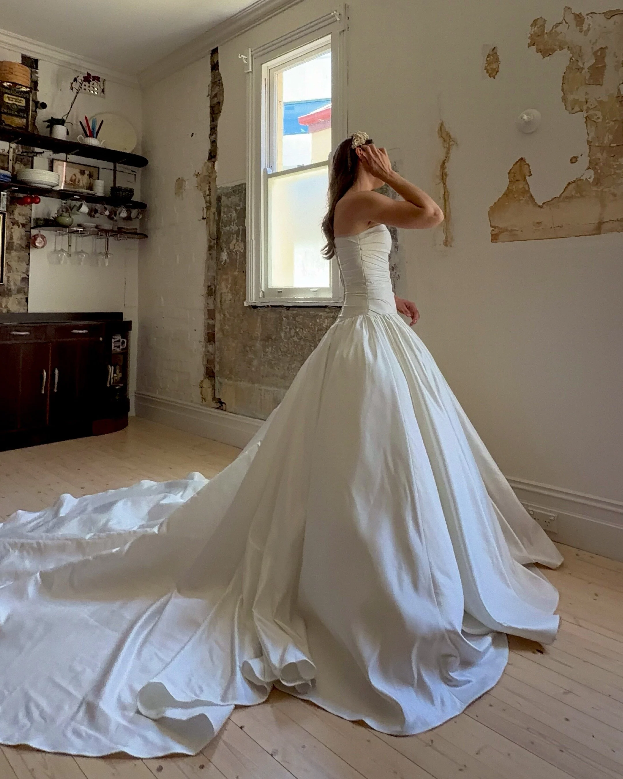 A woman in a white dress standing near an open window in a room with partially exposed walls, with her hand touching her head and a large piece of white fabric or dress material on the floor.