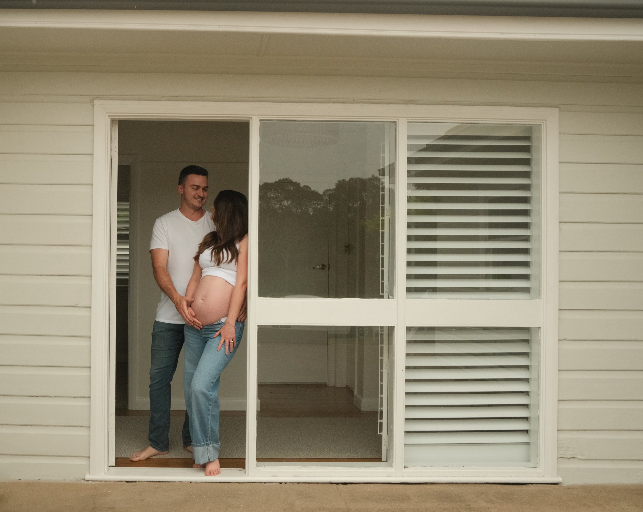 A pregnant woman and her partner standing in a doorway, smiling and holding her belly inside a house.