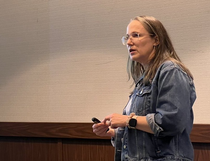 A woman with long hair, glasses, and a denim jacket giving a keynote address in front of a light-colored wall.