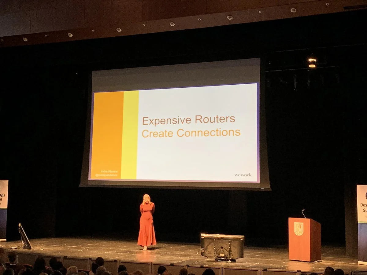A woman in a red dress standing on stage in front of a large screen that reads 'Expensive Routers Create Connections' at a conference.