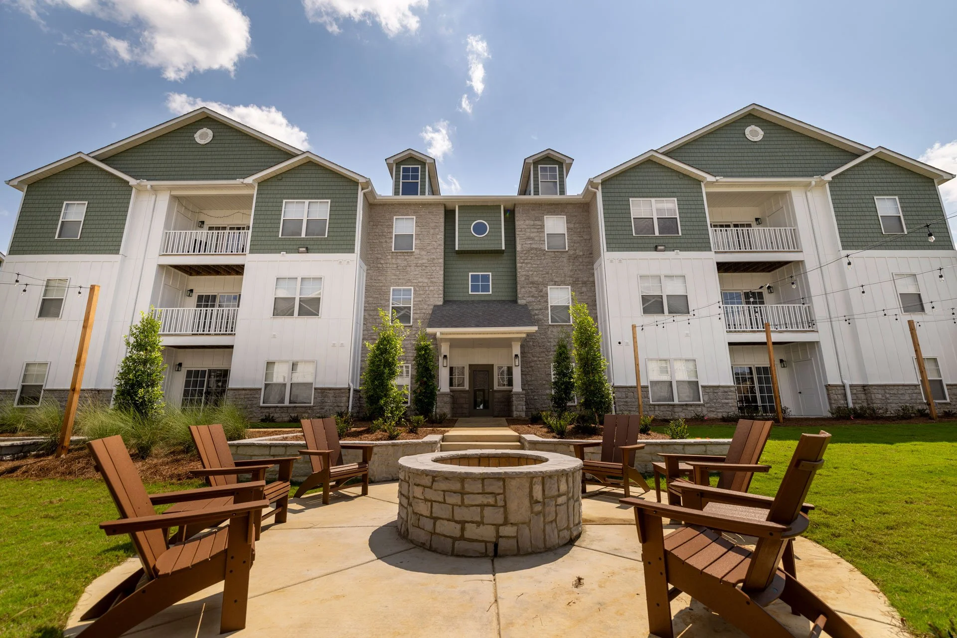 A modern multi-story condo building with green and white exterior, balconies, and a central entrance surrounded by landscaped greenery and a circular fire pit with wooden chairs in a courtyard.