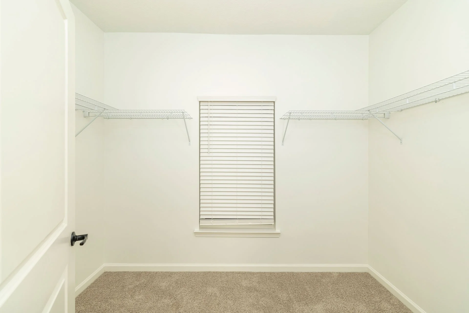 Empty walk-in closet with white walls, beige carpet, a single window with closed blinds, and two white wire shelves on each side.