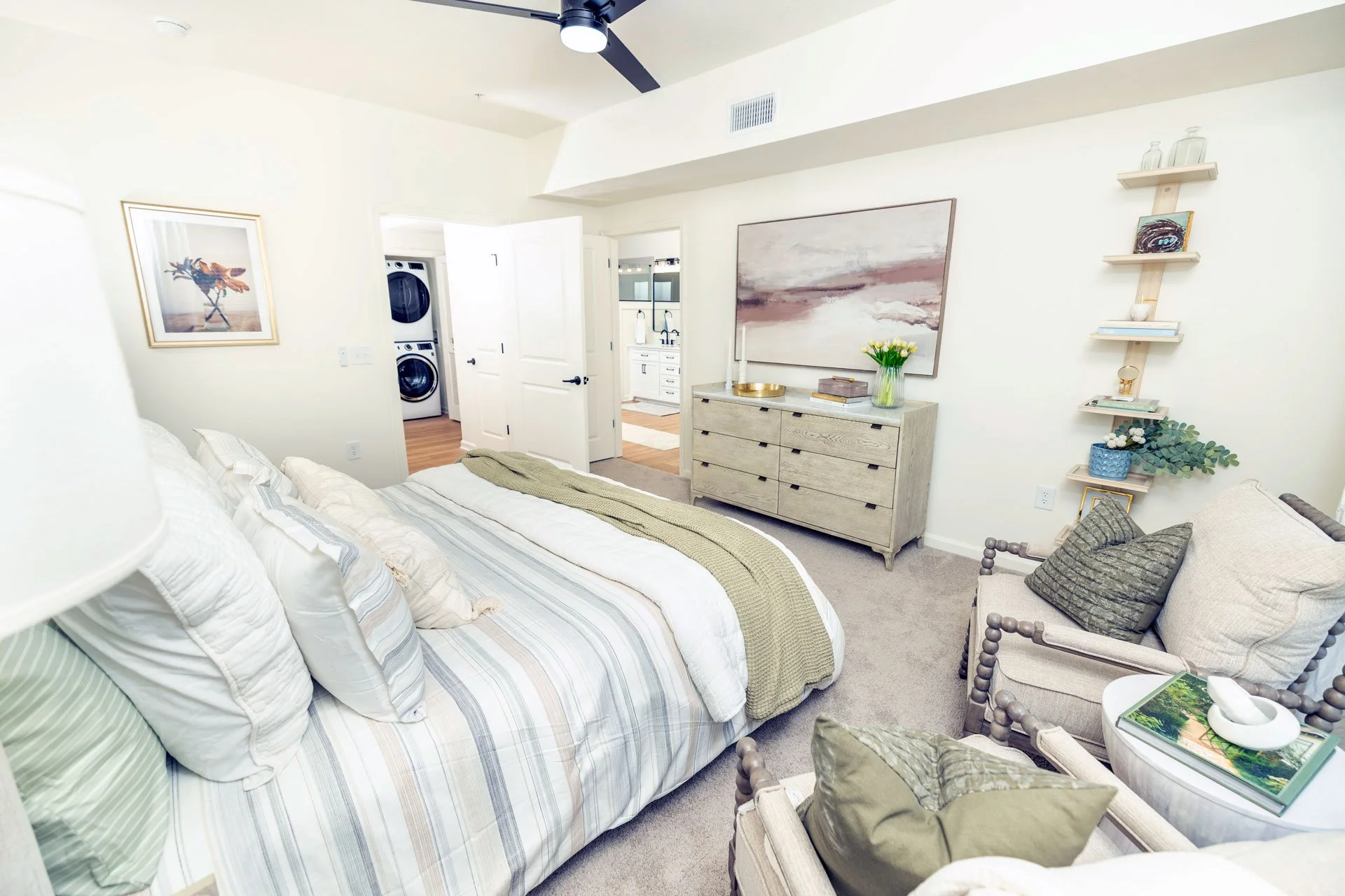 A cozy bedroom with a neatly made bed with striped bedding and multiple pillows. There are two armchairs with cushions, a small side table with magazines and a decorative item, and a white dresser with a large abstract painting above it. Shelves hold