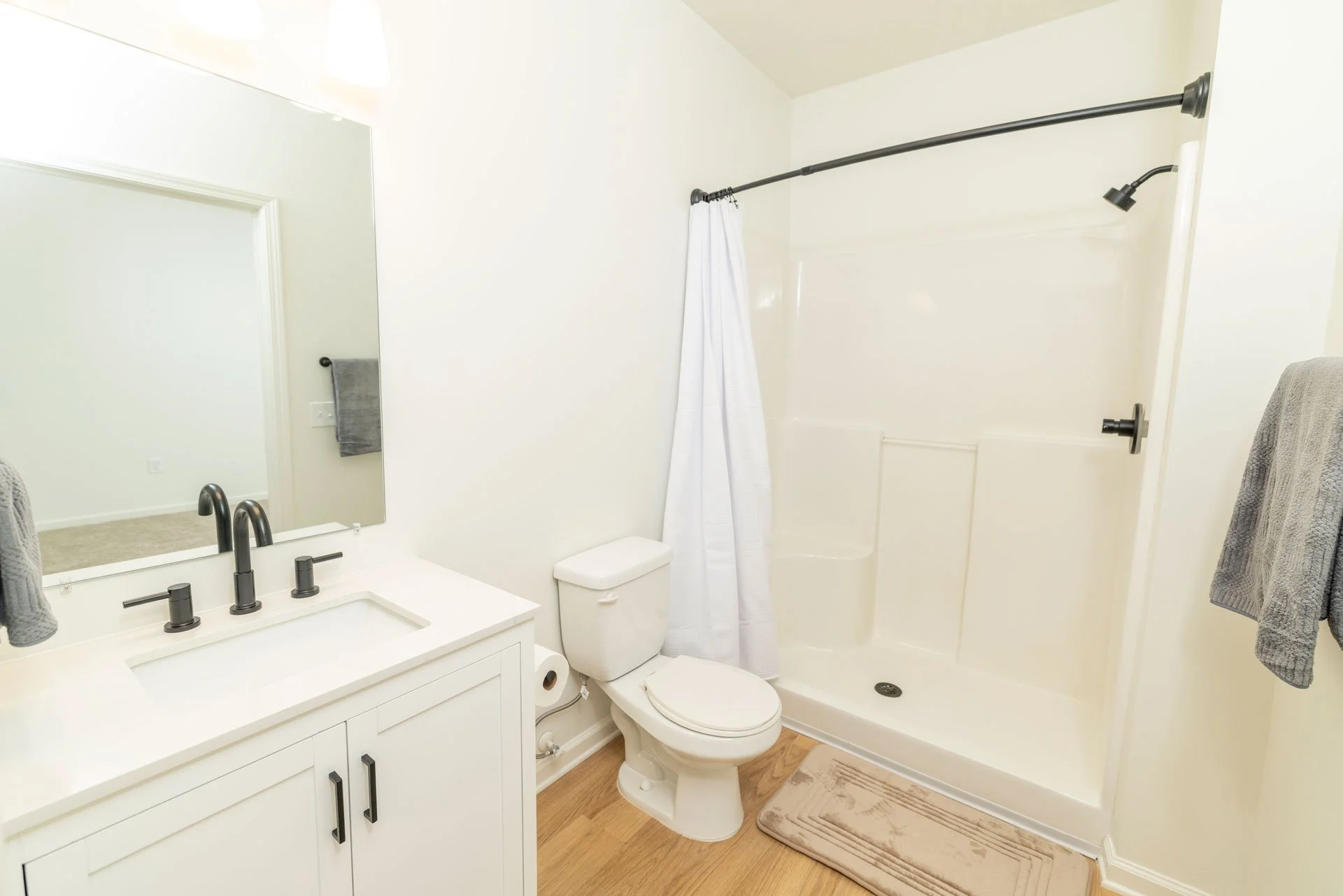 A clean, minimal bathroom featuring a white vanity with a black faucet, a mirror, a toilet, and a shower with a white curtain and black fixtures, alongside gray towels and a beige bath mat on wooden flooring.