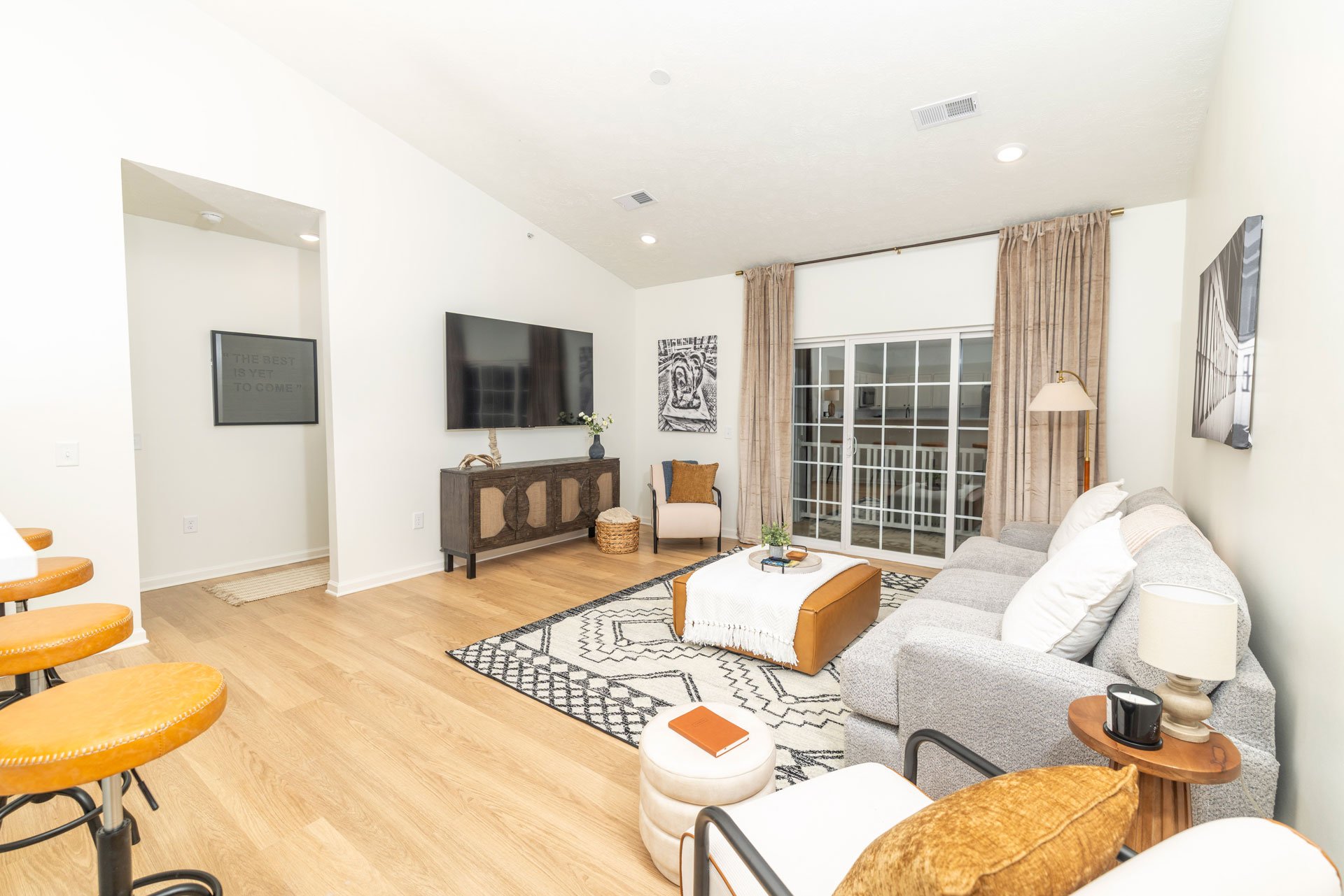 Living room with a gray sofa, white pillows, wooden side table with lamp, patterned rug, sliding glass door with beige curtains, wall-mounted TV, artwork, armchair, coffee table, bar stools.