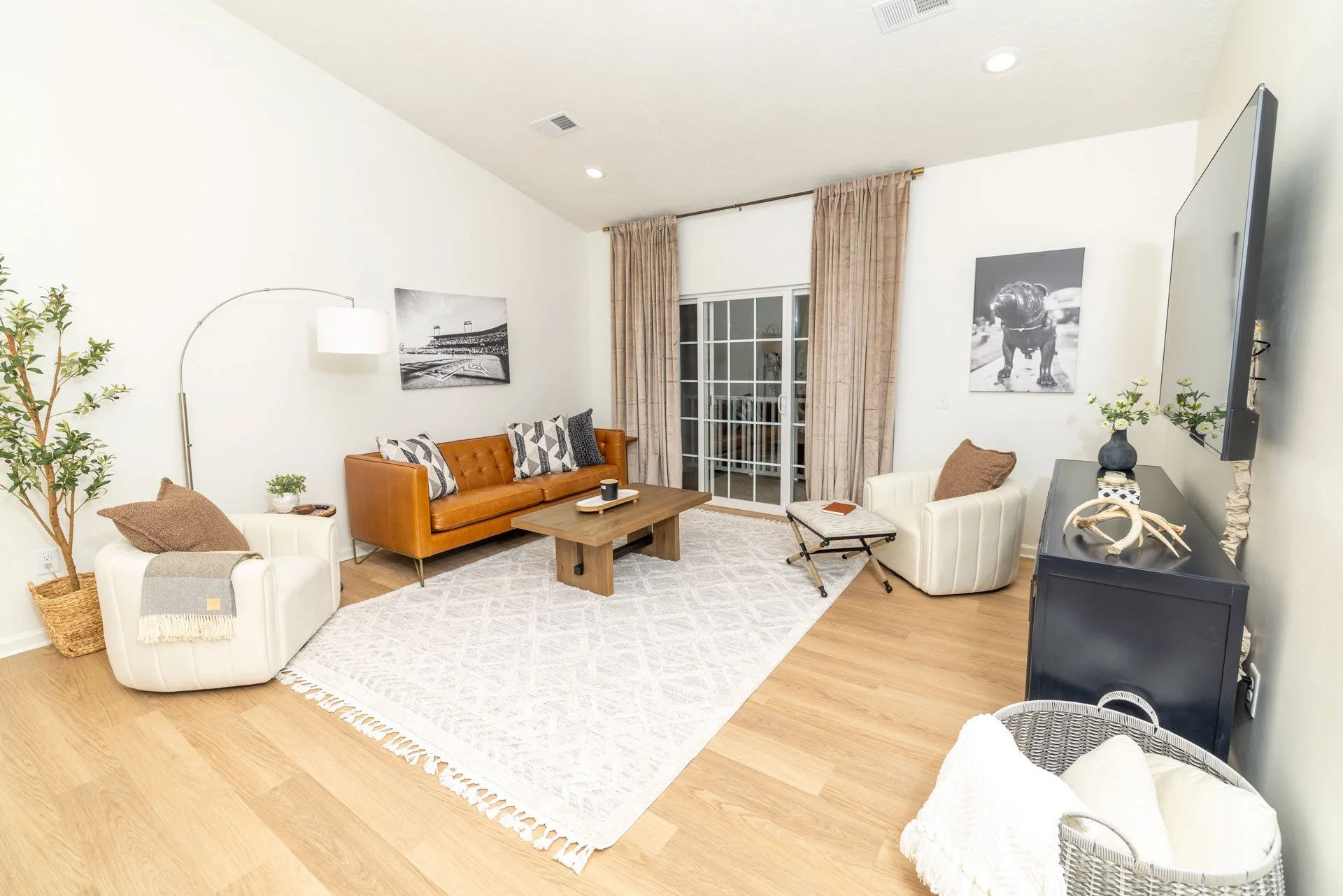 Living room with wooden flooring, a large area rug, a brown leather sofa, white armchairs with brown pillows, a wooden coffee table, a sliding glass door with beige curtains, wall art, a black media cabinet, a television, a vase with flowers, and dec
