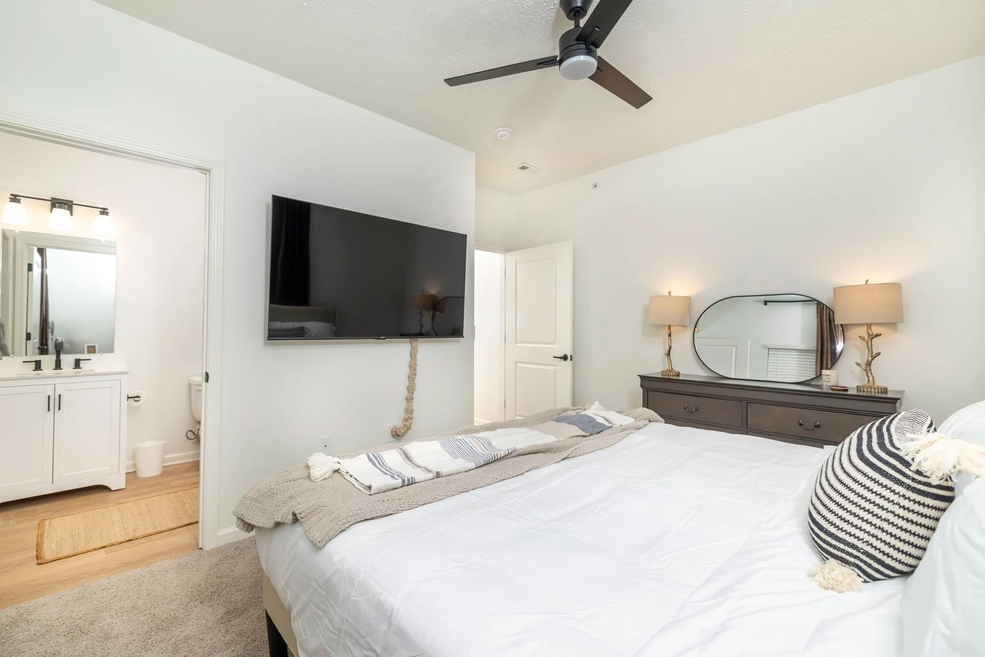 A bedroom with a bed, dresser, wall mirror, two lamps, a mounted TV, and a ceiling fan. The bed has a striped pillow and a towel.