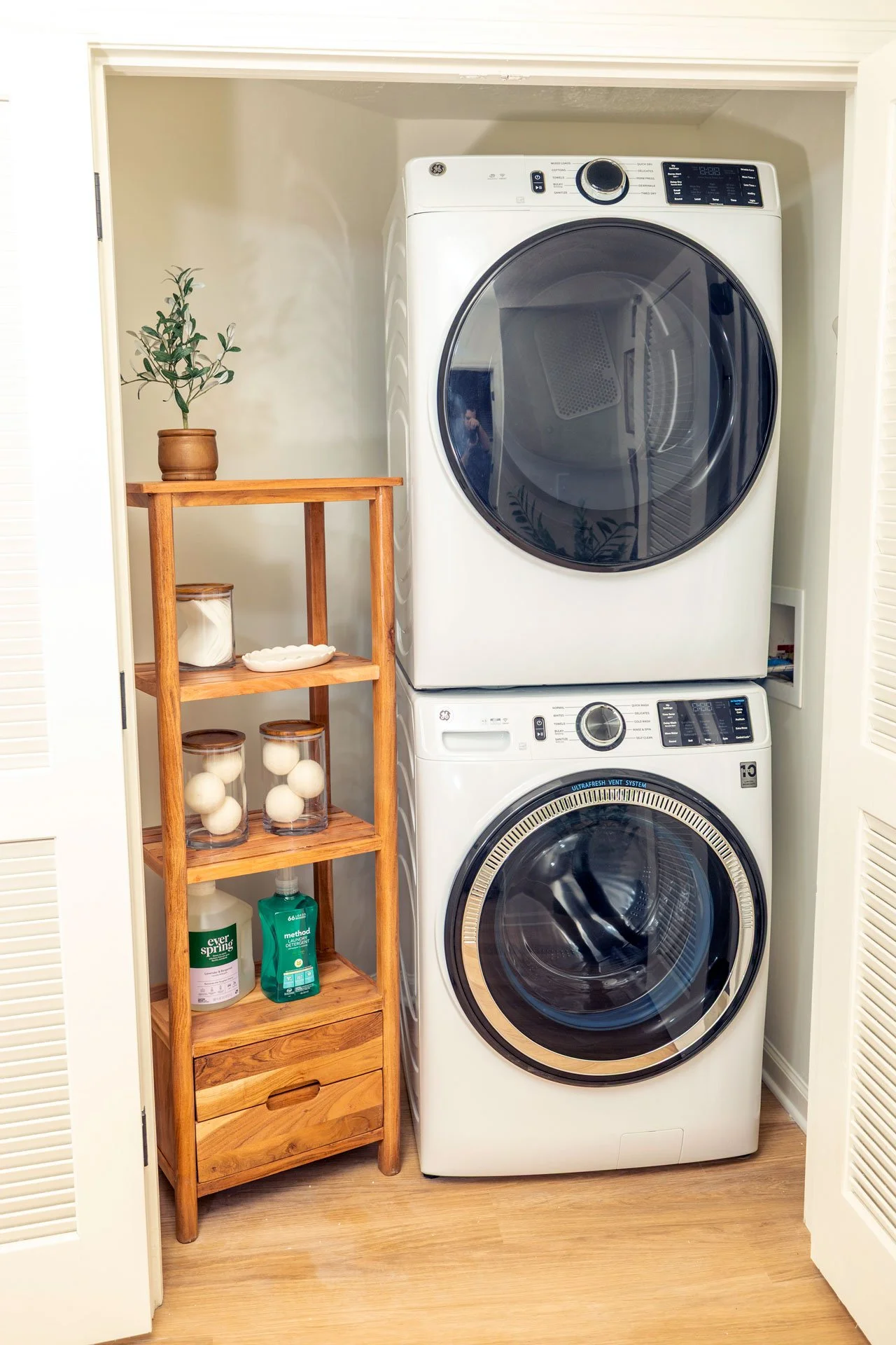 A stacked white front-loading washer and dryer in a laundry closet with a wooden shelf holding cleaning supplies and decorative items.