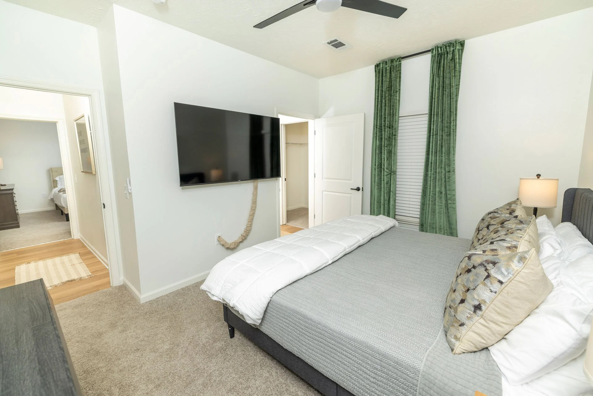 Hotel bedroom with bed, pillows, nightstand, lamp, green curtains, wall-mounted TV, ceiling fan, and open closet door