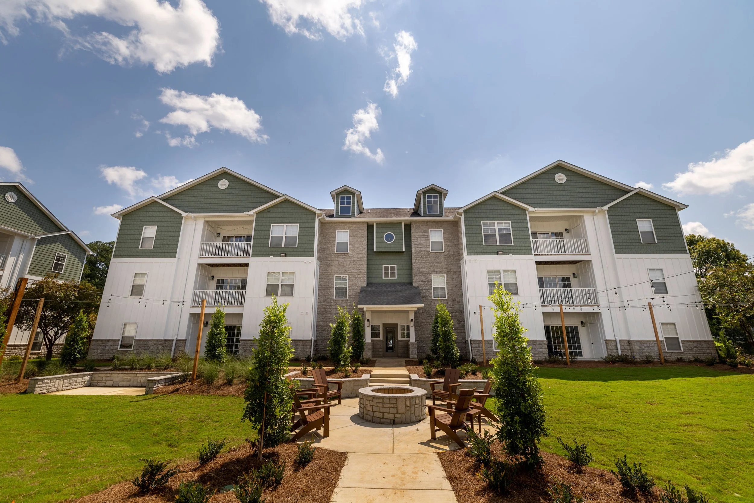 Front view of a multi-story apartment building with green and white exterior, small balconies, and a landscaped courtyard with chairs surrounding a fire pit.