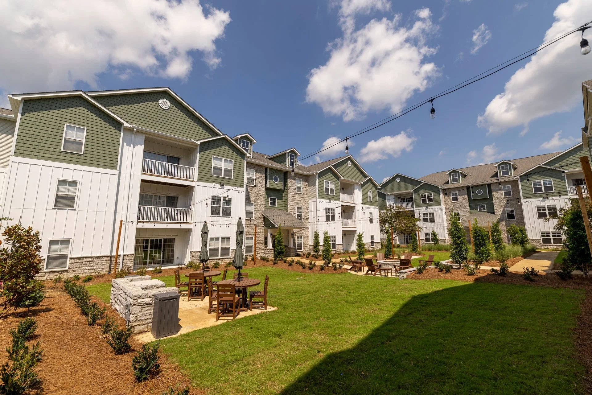 Outdoor courtyard with green grass, trees, and several wooden tables with umbrellas in front of multi-story apartment buildings with green and white exteriors, under a partly cloudy blue sky.