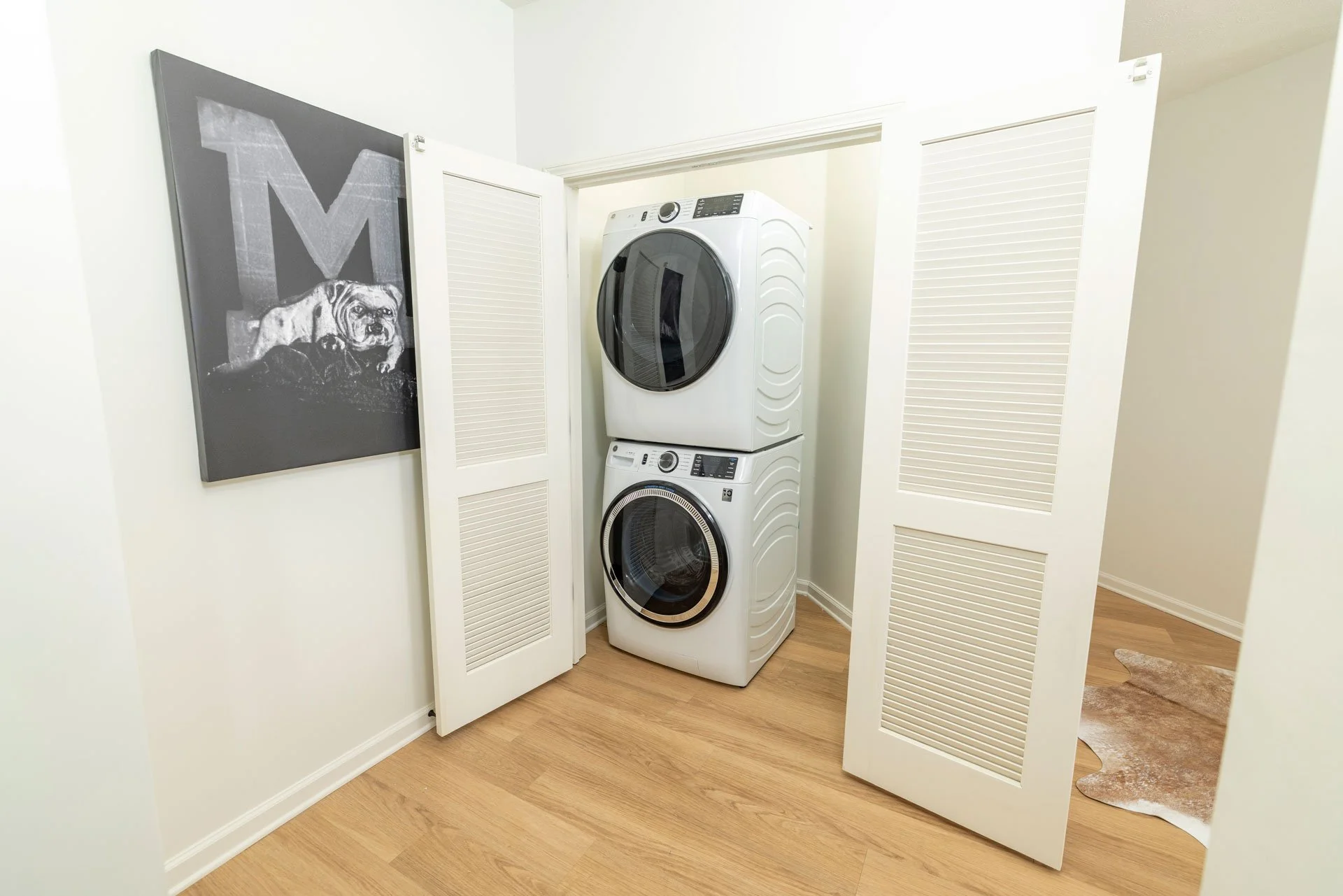 A laundry room with a stacked washing machine and dryer inside a closet with white louvered bifold doors, open to reveal the appliances and light wood floor, with a black and white bulldog painting on the wall.