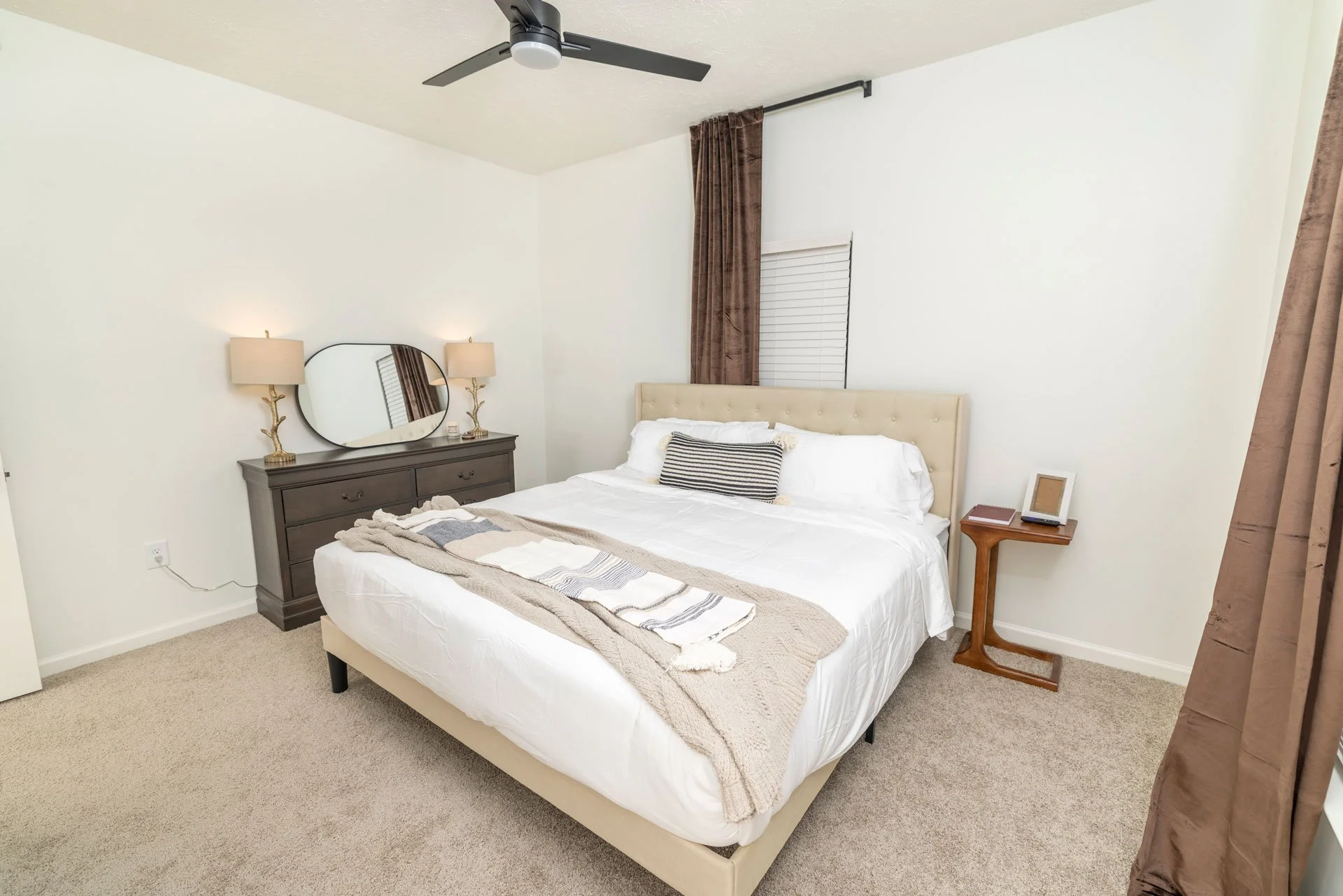 A bedroom with a bed, nightstand, dresser, mirror, lamps, window with blinds and curtains, ceiling fan, and beige carpet.