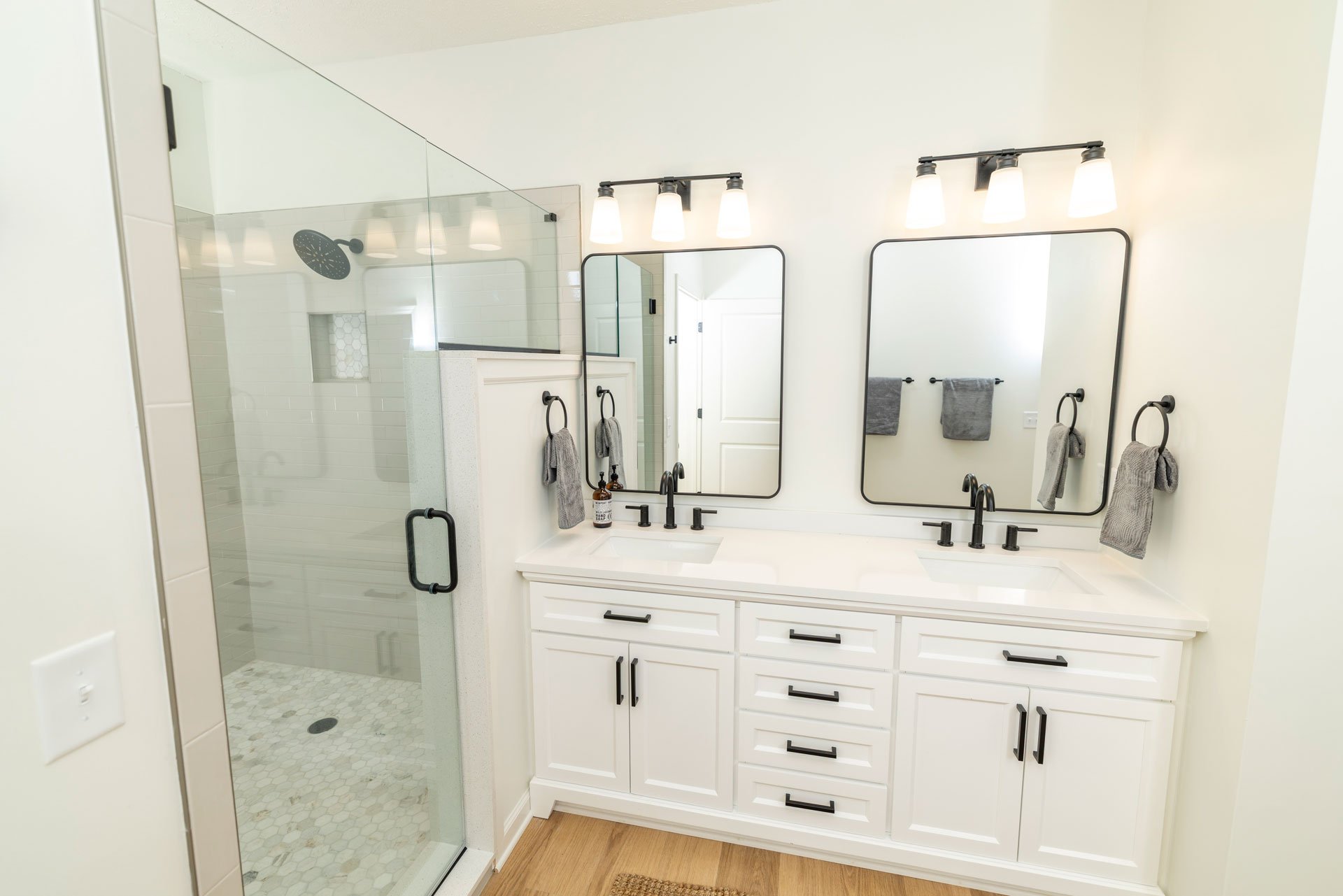 A modern bathroom with a glass-enclosed shower, white cabinetry with black hardware, two rectangular mirrors above the double vanity, and black light fixtures. Gray towels hang on rings next to each mirror.