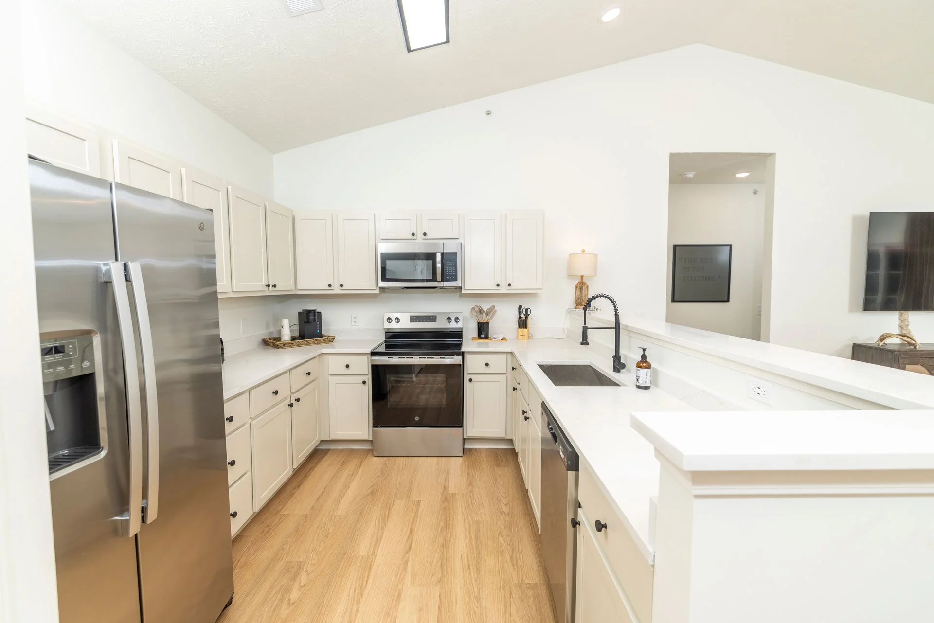Kitchen with white cabinets, stainless steel refrigerator, oven, microwave, black sink, and wooden flooring.