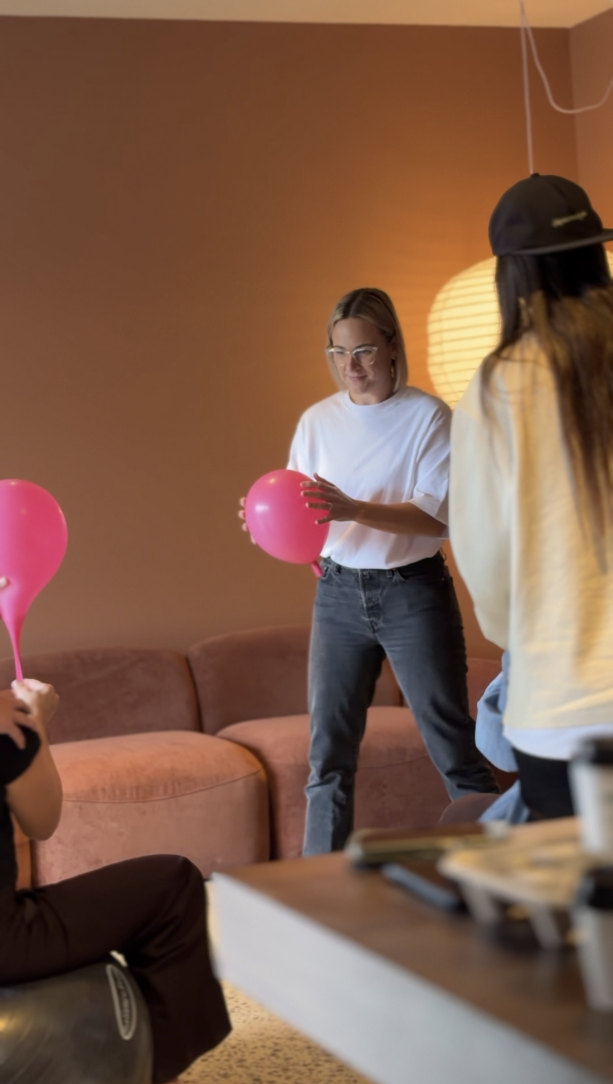 People holding pink balloons in a room with a brown couch and a warm-colored wall, with some standing and others sitting.