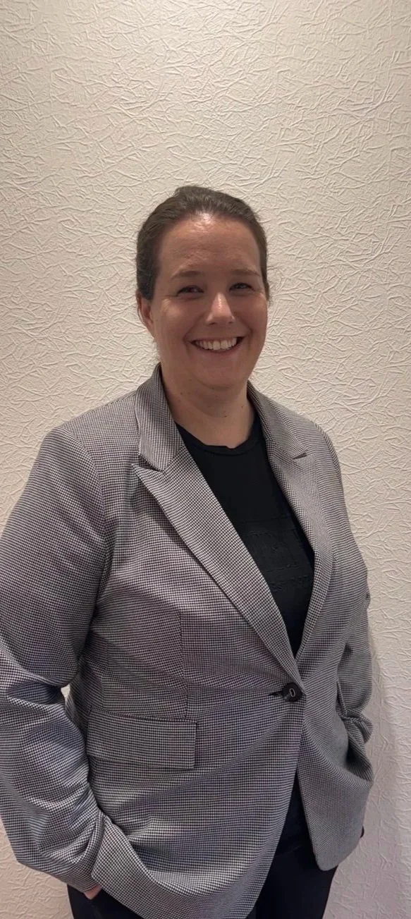 A Clinical Psychologist with brown hair in a bun, wearing a gray blazer over a black shirt, smiling, standing against a textured beige wall.
