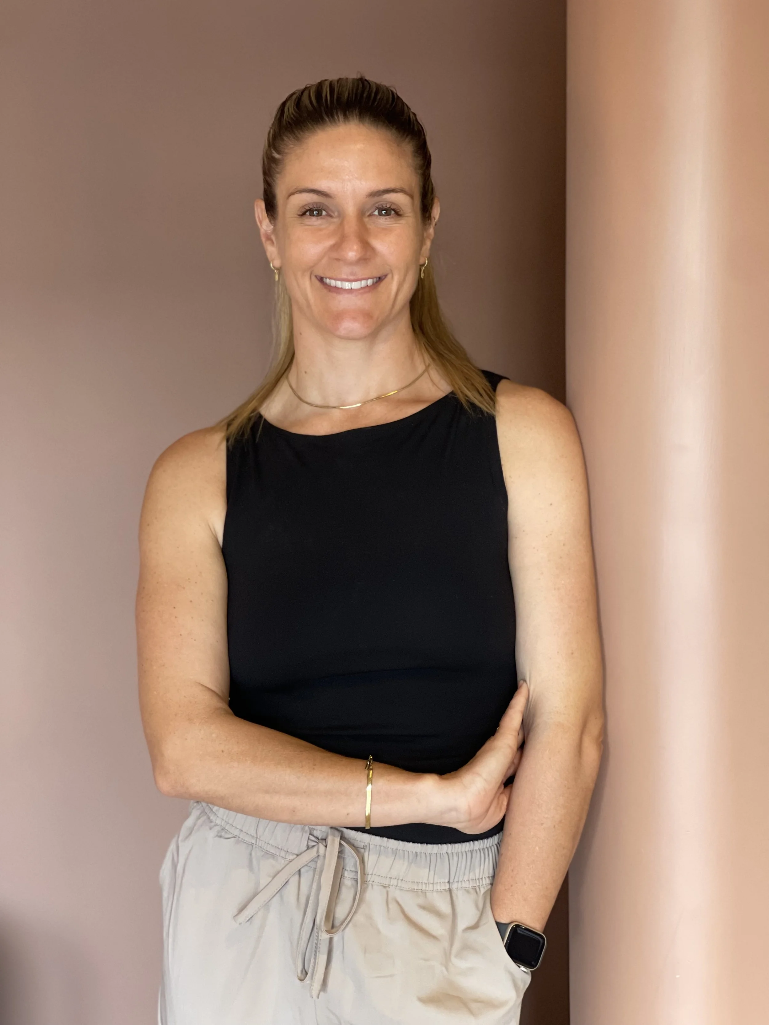 A woman with light brown hair tied back, wearing a black sleeveless top, beige drawstring shorts, a bracelet, and an Apple Watch, standing against a two-tone pink wall.