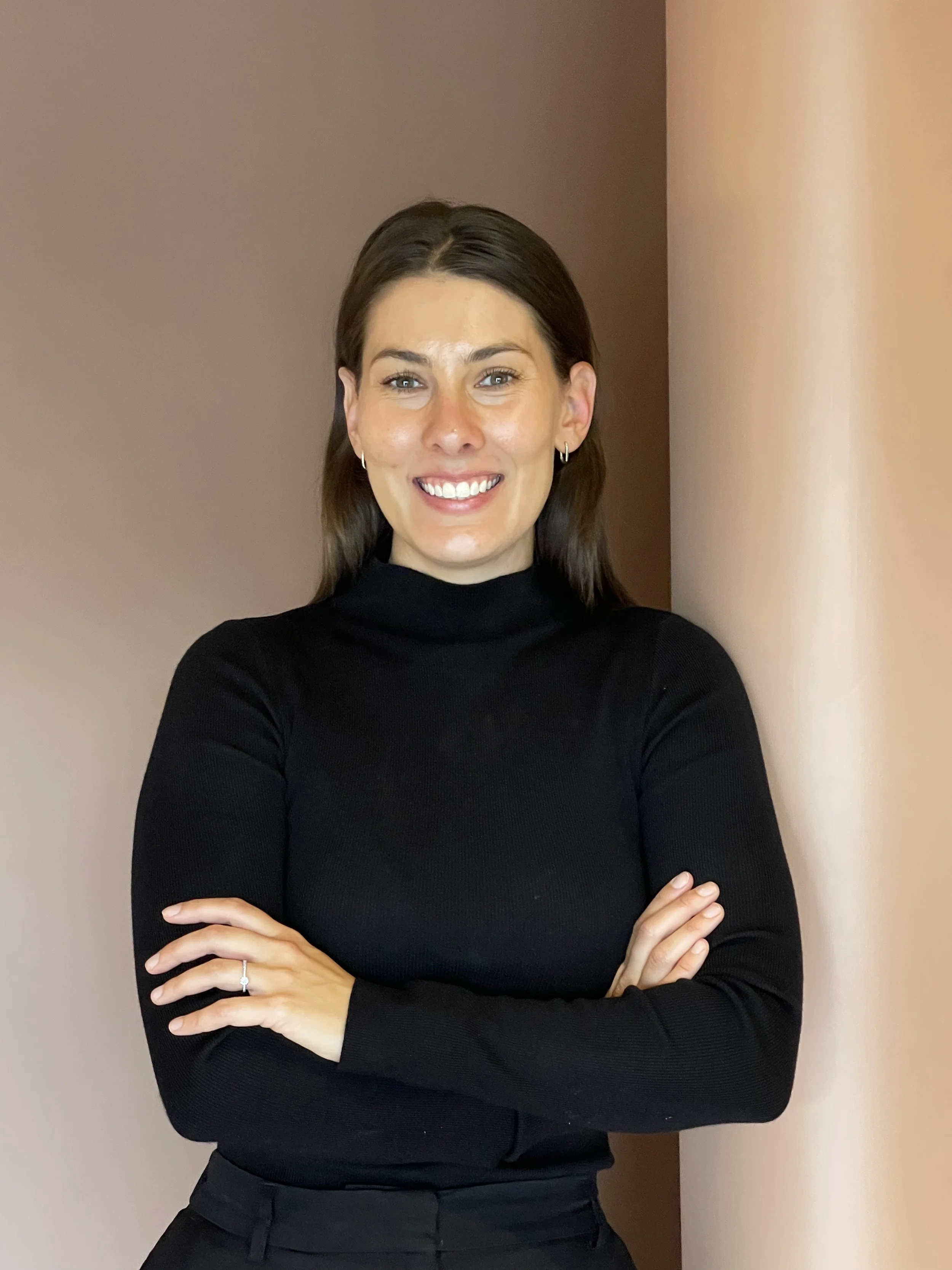 A woman with brown hair, light skin, and blue eyes, smiling, standing with arms crossed, wearing a black turtleneck top, against a beige background.