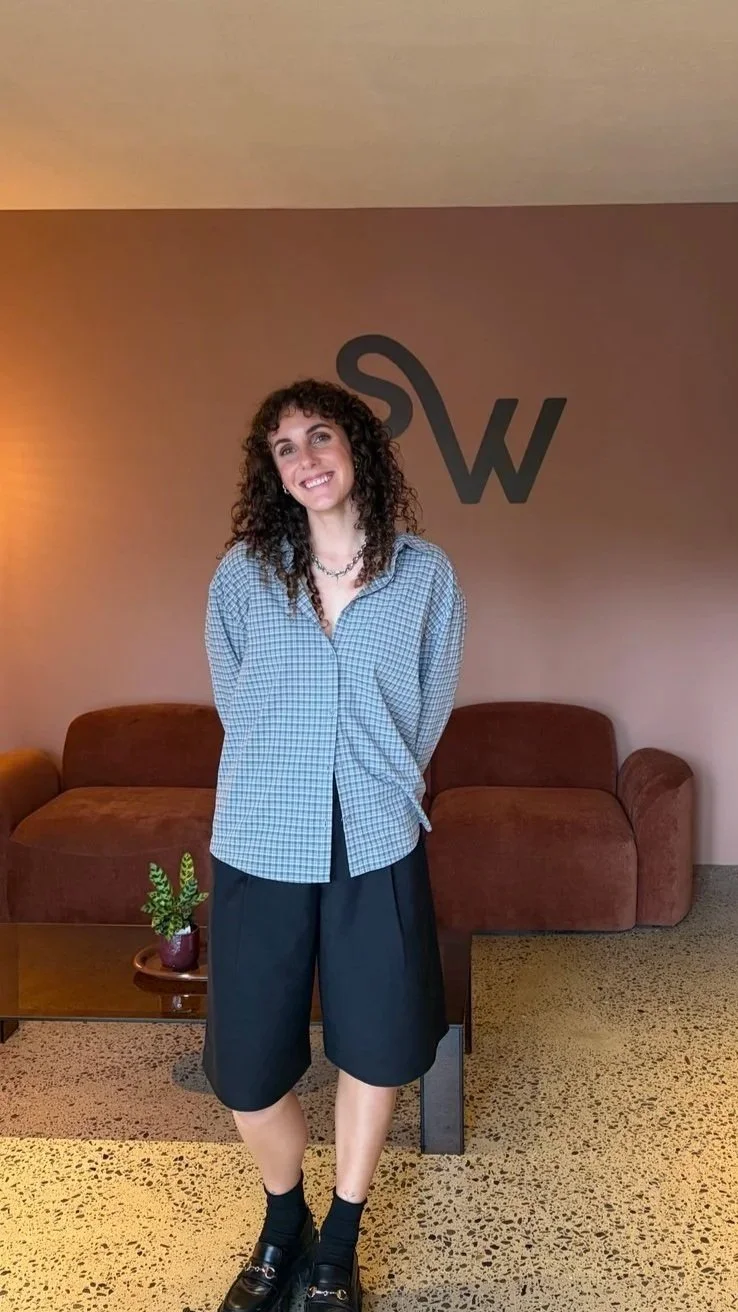 A woman with curly dark hair smiling, standing in front of a wall with a large letter 'W' on it. She is wearing a blue checkered shirt, black wide-leg shorts, black socks, and black shoes. There's a brown tufted sofa and a small table with a potted plant in the background.