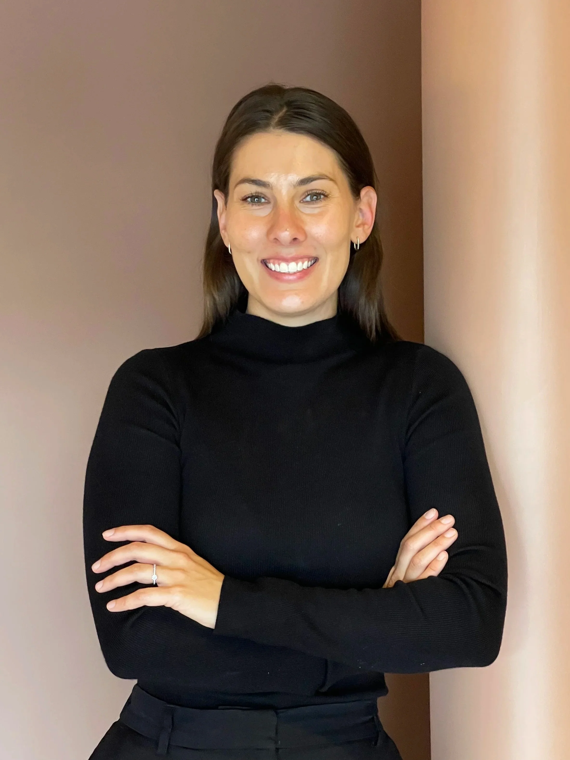 A woman with dark brown hair wearing a black turtleneck and black pants, standing against a neutral background with her arms crossed, smiling and looking at the camera.