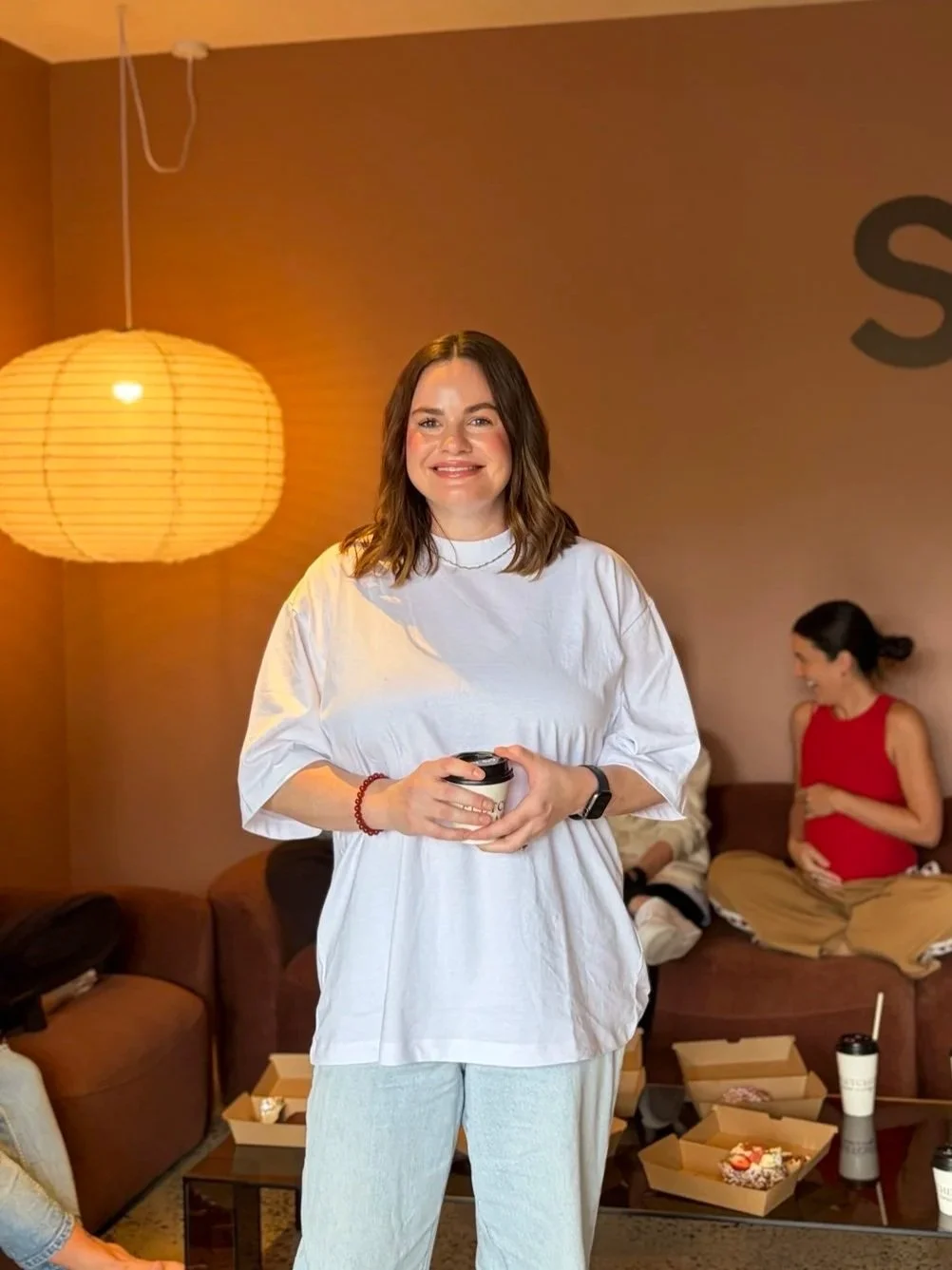 A woman with shoulder-length brown hair holding a cup, smiling, in a cozy room with a warm-colored wall and a large paper lantern hanging from the ceiling. In the background, a pregnant woman in a red top and beige pants sitting on a sofa, with snacks and drinks on the table.