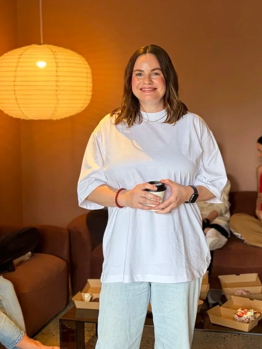 A woman smiling while holding a coffee cup, standing in a cozy living room with warm lighting, a paper lantern lamp, and people in the background sitting on couches.